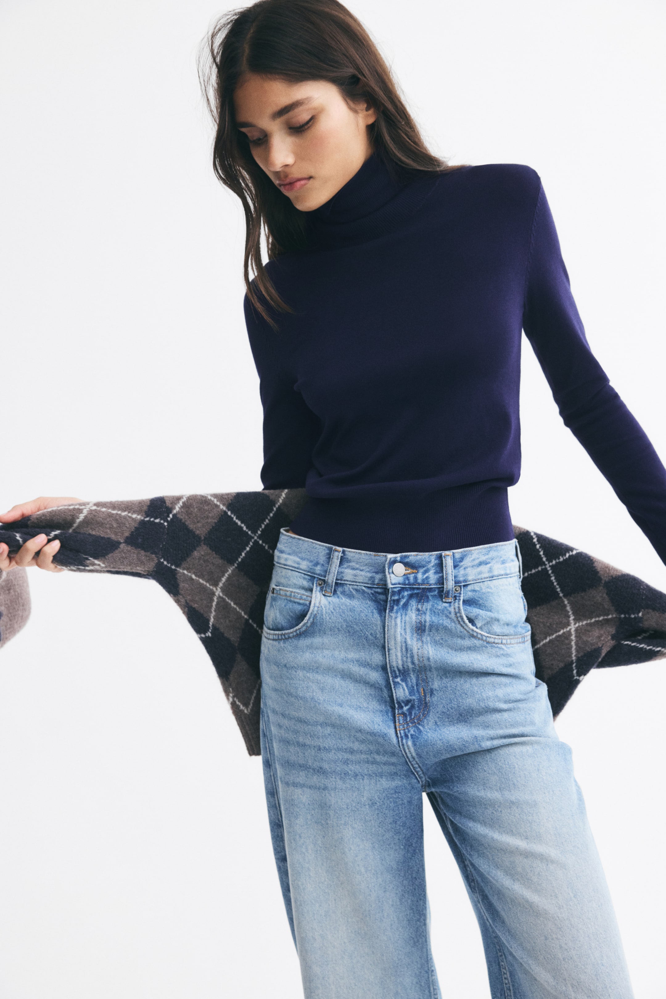 View larger image: A navy blue, long-sleeved turtleneck sweater is being worn by the model, tucked into high-waisted, light-wash blue jeans. A brown and navy argyle-patterned knit cardigan is being held behind the waist by the model.