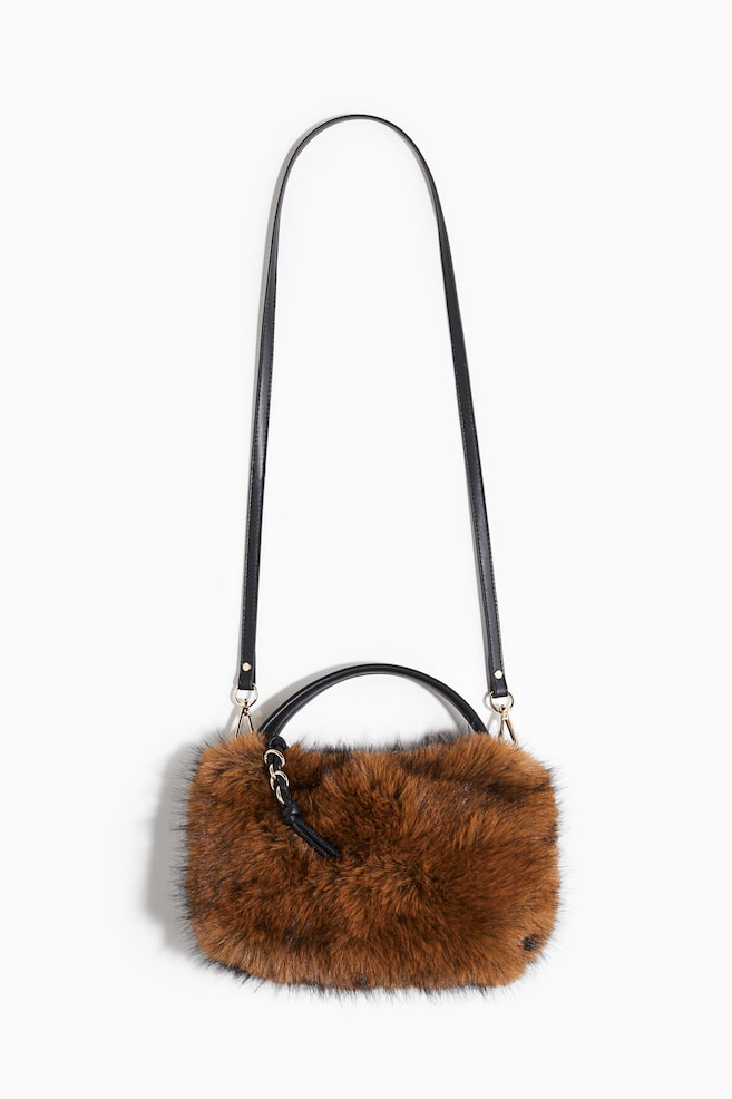 Un sac à main en fausse fourrure marron et noire est suspendu, présentant son devant, avec une poignée supérieure noire et une longue bandoulière noire fine attachée avec des ferrures dorées. Une languette noire avec un détail de chaîne dorée est visible à l'avant. - 1