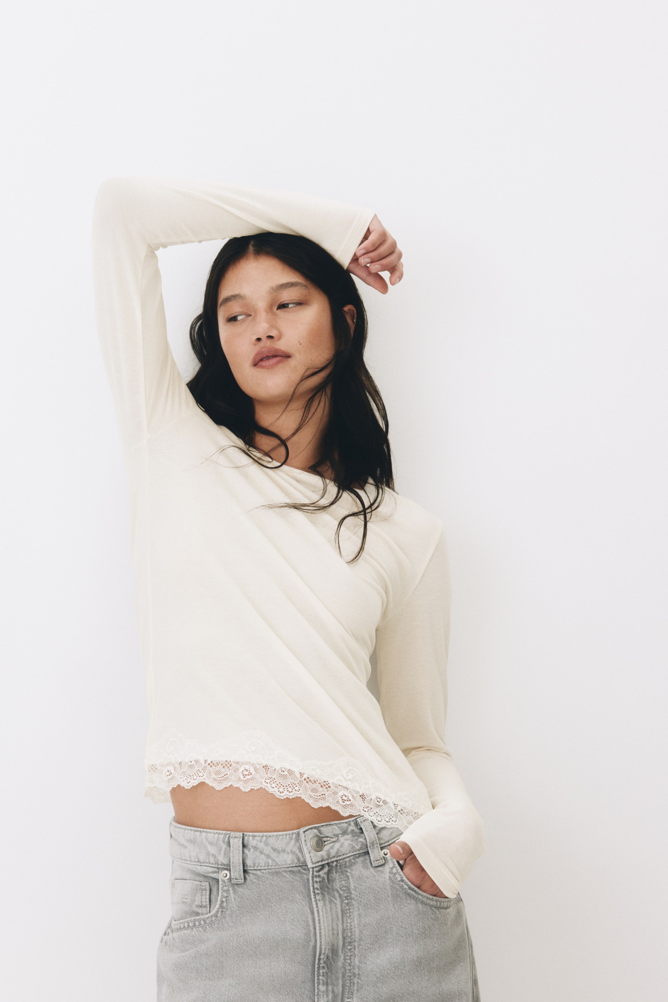 View larger image: The model is donning a snug, long-sleeved top crafted from soft jersey, featuring a round neckline and a hem adorned with lace. She raises one arm, showcasing the stretch of the sleeve, while her other hand slips into the pocket of her light grey jeans.