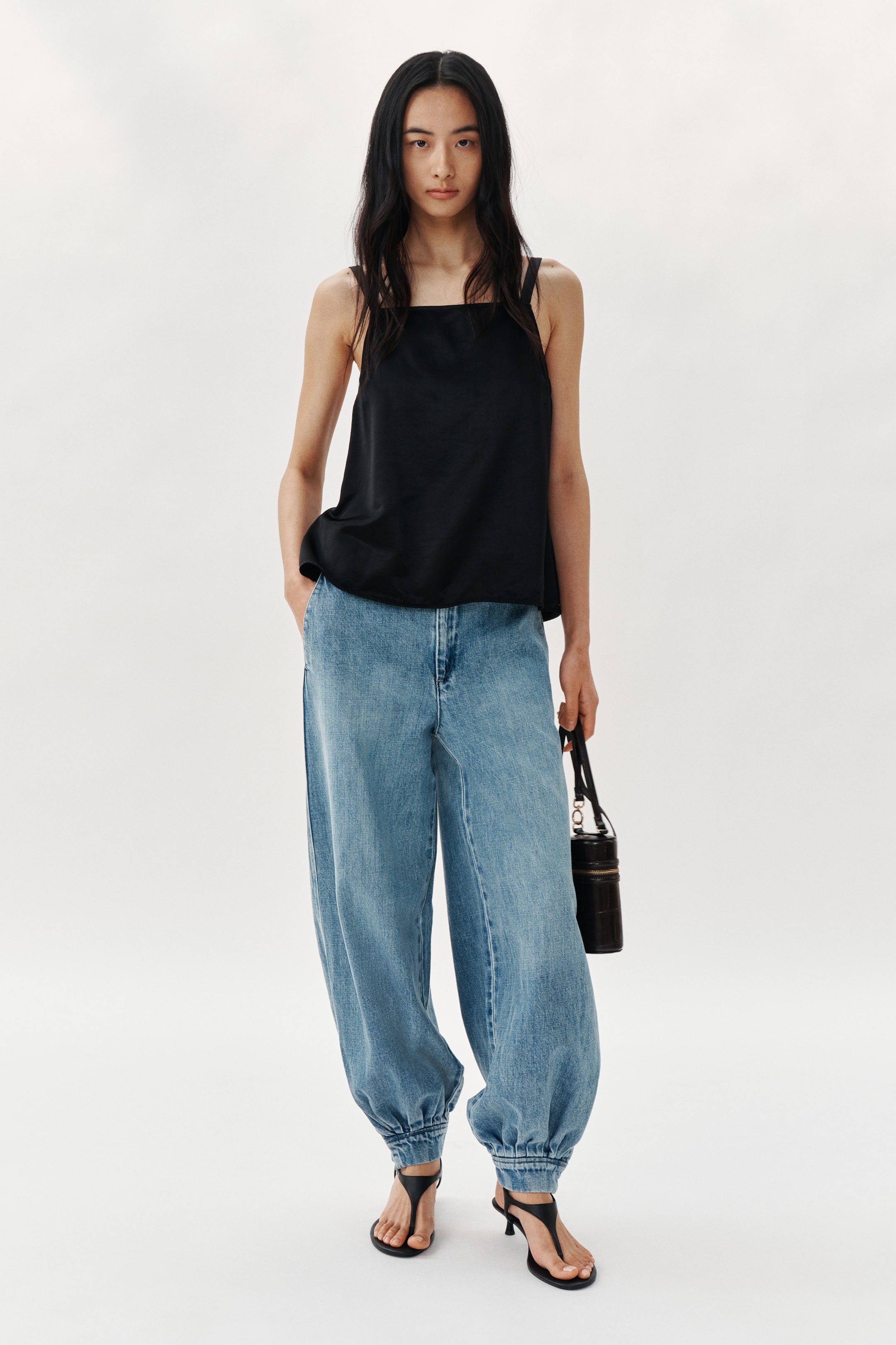 View larger image: A black square-neck top na may malapad na straps at maluwag na fit, pinapartneran ng light wash na balloon-leg jeans na may cinched na bukong-bukong. Ang black thong sandals na may kitten heel ay kumukumpleto sa look, at hawak ang isang black cylindrical bag.
