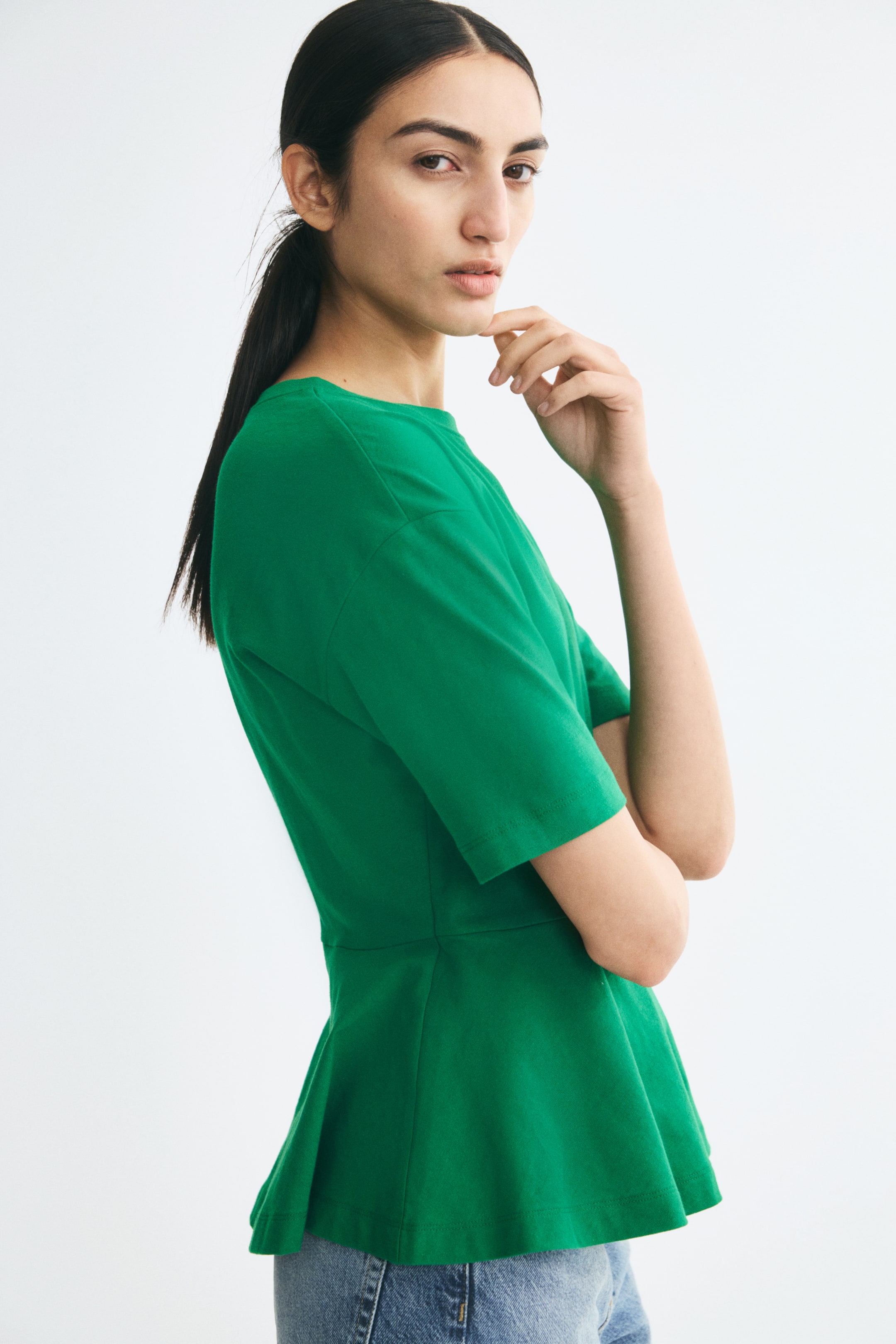 View larger image: The model is wearing a green cotton jersey top featuring a round neckline, dropped shoulders, short sleeves, and a flared peplum hem. Standing in profile with arms folded, the model styles the top with light blue denim jeans.