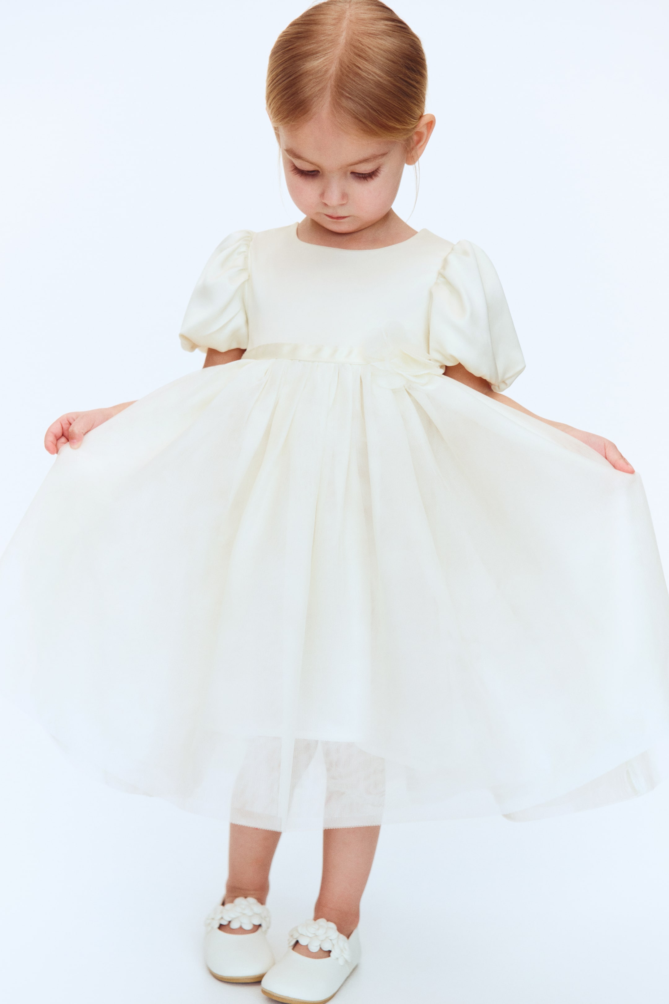 Größeres Bild ansehen: Ein elfenbeinfarbenes Partykleid mit einem glatten Oberteil und Puffärmeln, einem runden Ausschnitt und einem vollen, plissierten Rock aus transparentem Stoff, der mit einem Satinband in der Taille umschlungen ist und seitlich zu einer Schleife gebunden wird. Weiße Mary Jane Schuhe mit Blumenverzierungen vervollständigen den Look.