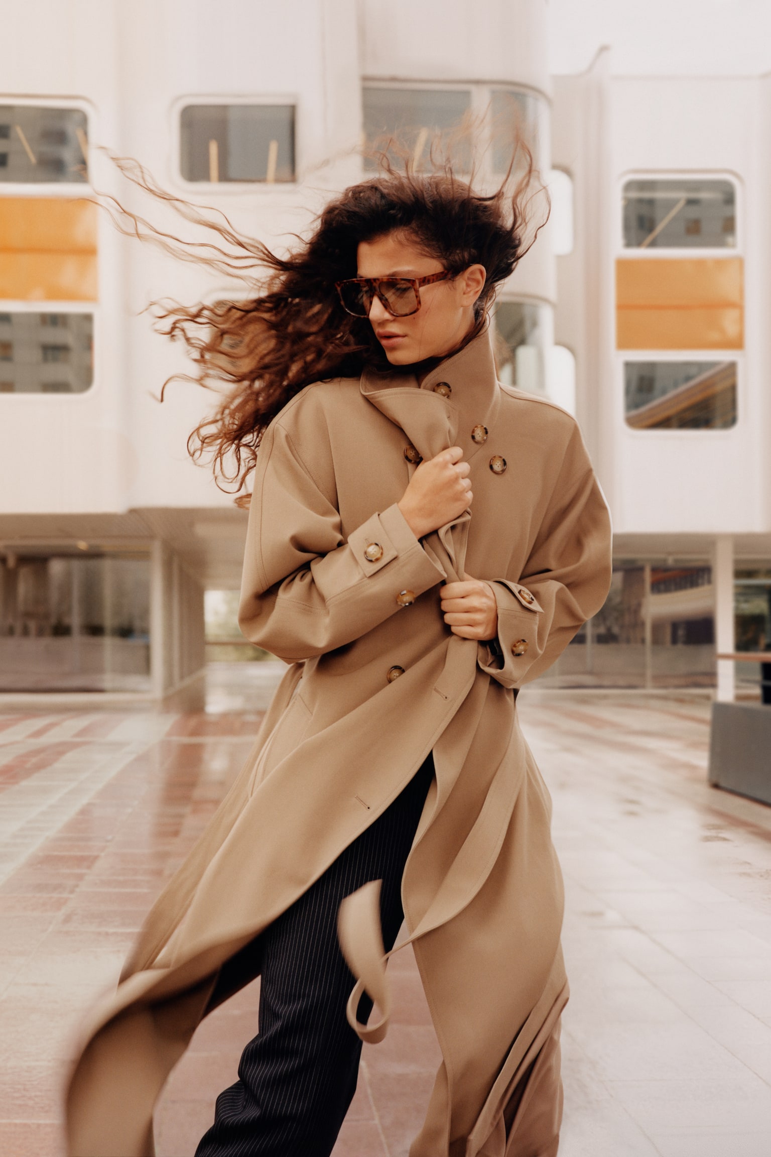 An oversized beige trench coat featuring a high collar, double-breasted button closure, and buttoned cuffs is being worn by the model. The model is holding the collar closed with both hands, styling the coat with dark pinstriped trousers. - 1