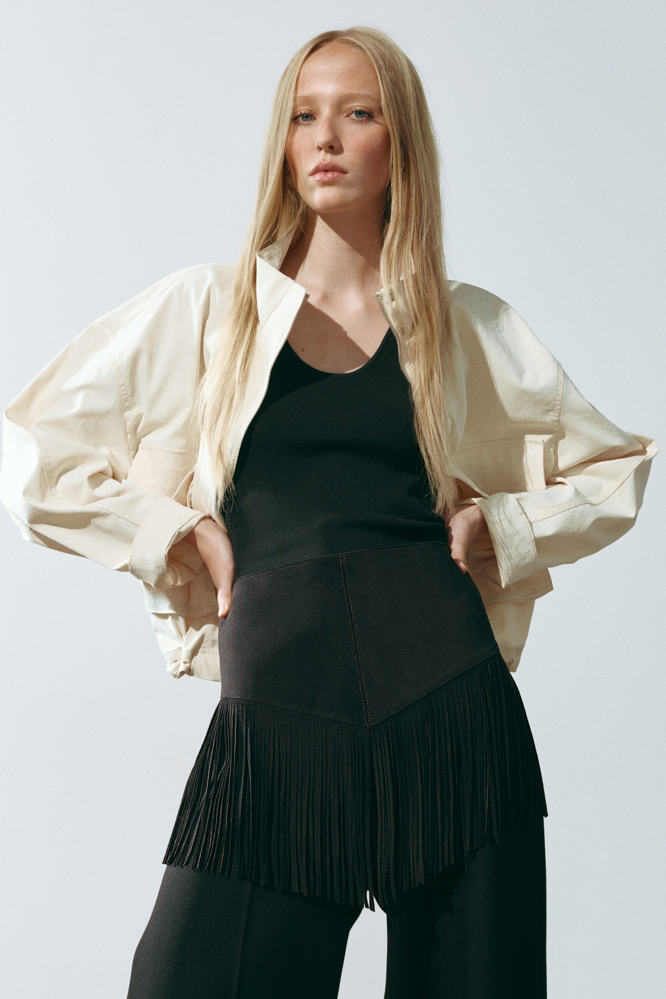 View larger image: A black belt in a napped-finish fabric is worn at the model’s waist, featuring a wide, downward-pointing triangular panel with long, dense fringe along the bottom edge. The belt is layered over a black top and matching trousers, creating a monochromatic base styled under an open cream-coloured jacket.