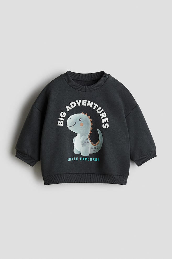 A dark gray sweatshirt is being shown from the front, featuring a ribbed crew neck, ribbed cuffs, and a ribbed hem. Two snap buttons are visible on the left shoulder, and a light blue cartoon dinosaur motif is centered on the front, with 'BIG ADVENTURES' arcing above it in white text and 'LITTLE EXPLORER' below it in light blue text. - 1