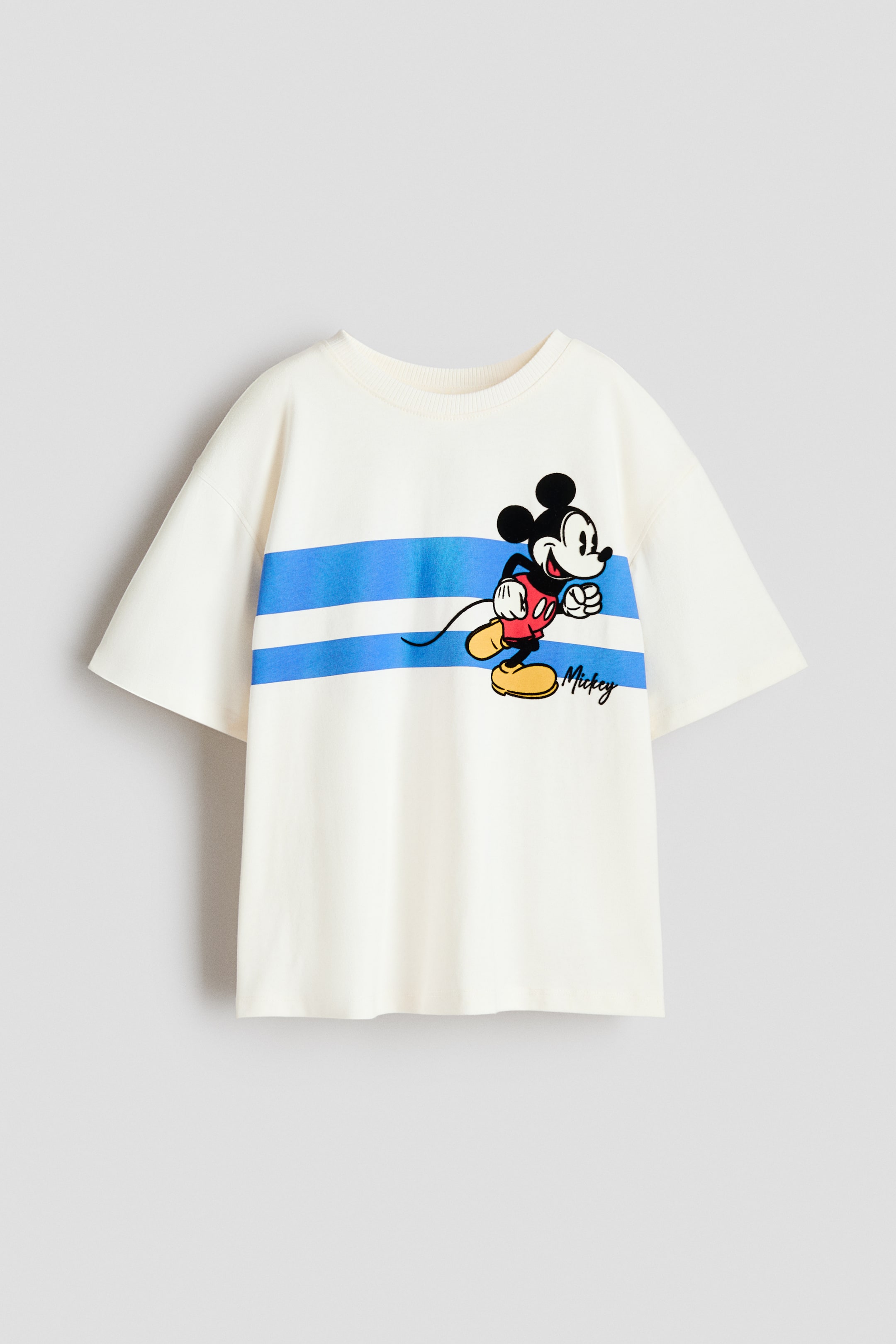 Ver imagen más grande: Una camiseta de gran tamaño en color crema se ve desde el frente, con un estampado de Mickey Mouse corriendo a través de dos franjas horizontales azules en el pecho. Está diseñada con un cuello redondo ribeteado y hombros caídos.