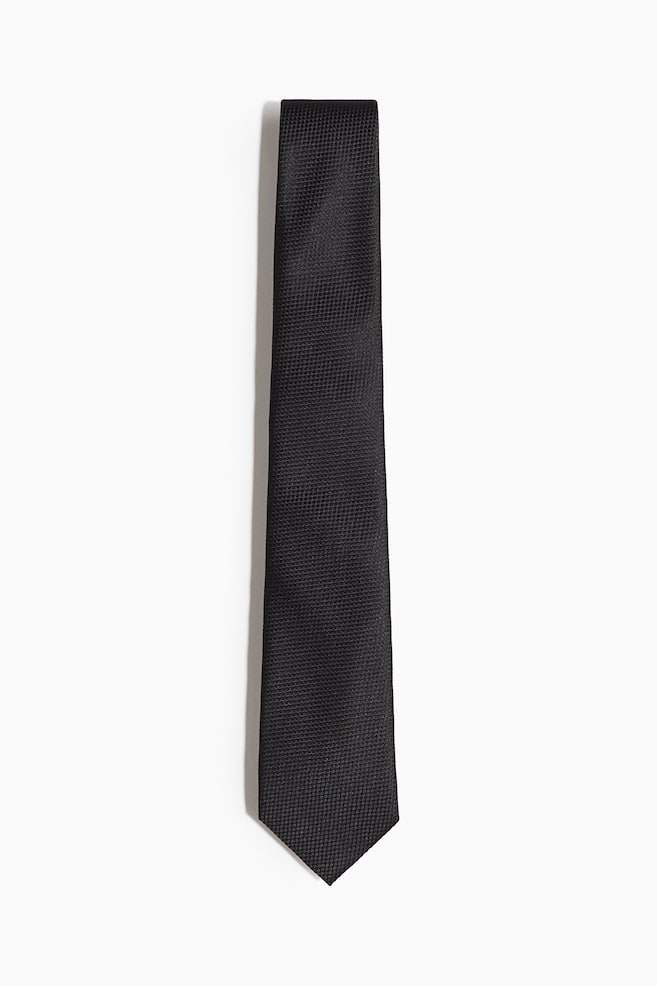 A classic black tie is presenting its front side, lying flat. It is featuring a visible textured weave across its entire surface and is tapering to a pointed bottom. - 1