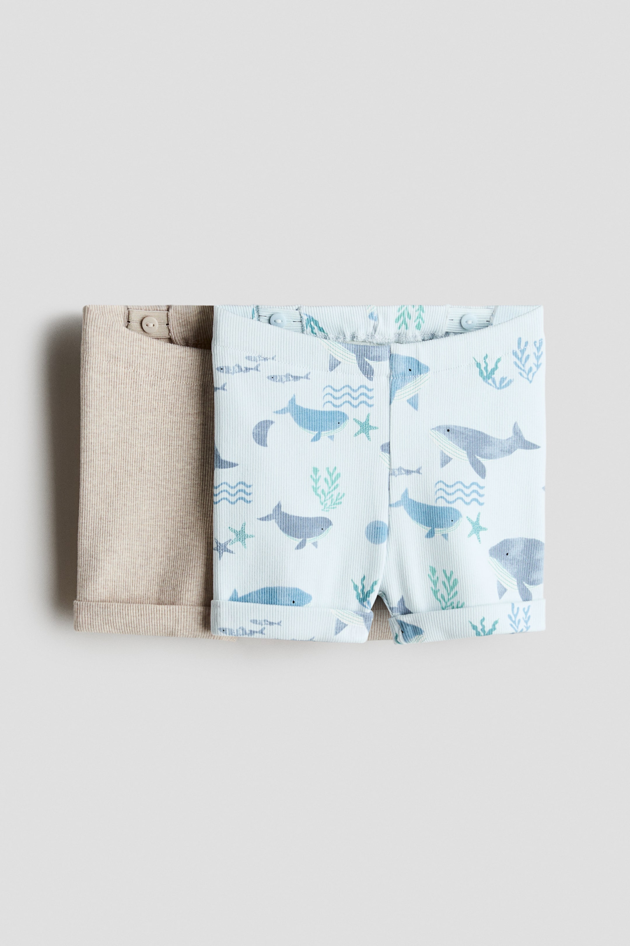 Grösseres Bild ansehen: Ein 2er-Pack Baumwoll-Jersey-Shorts wird frontal präsentiert, horizontal gefaltet. Ein Paar hat ein beige geripptes Design, während das andere ein hellblaues geripptes Design mit einem meeresthematischen Muster aus Walen, Fischen und Algen aufweist; beide Shorts zeigen elastische Bundabschlüsse mit sichtbaren Knöpfen und umgeschlagenen Säumen.