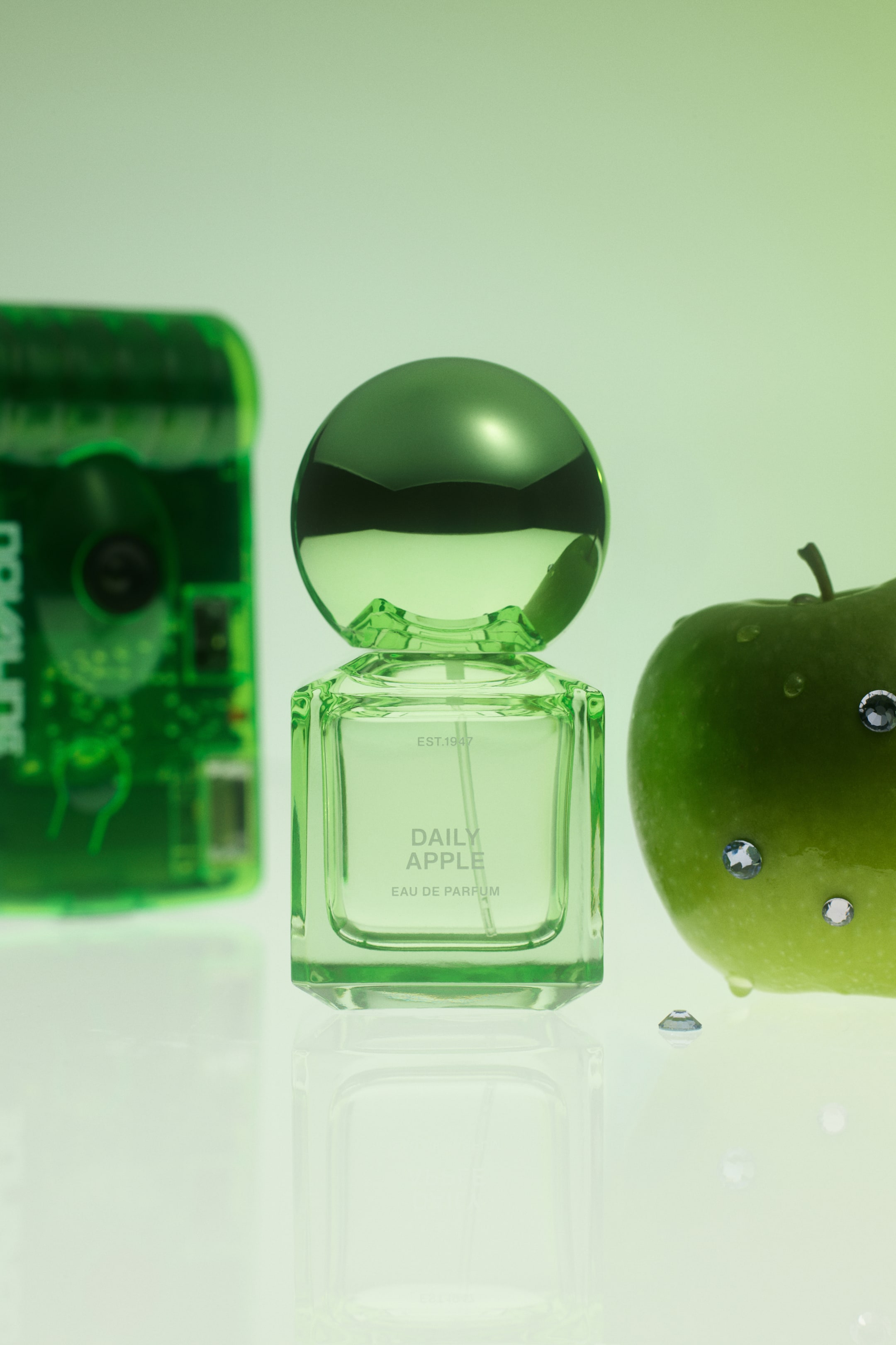 View larger image: A transparent green glass perfume bottle labelled "DAILY APPLE EAU DE PARFUM" is standing upright, featuring a square body and a large, metallic-finish spherical green cap. Resting on a reflective surface, the bottle is positioned beside a fresh green apple adorned with small, sparkling crystals.