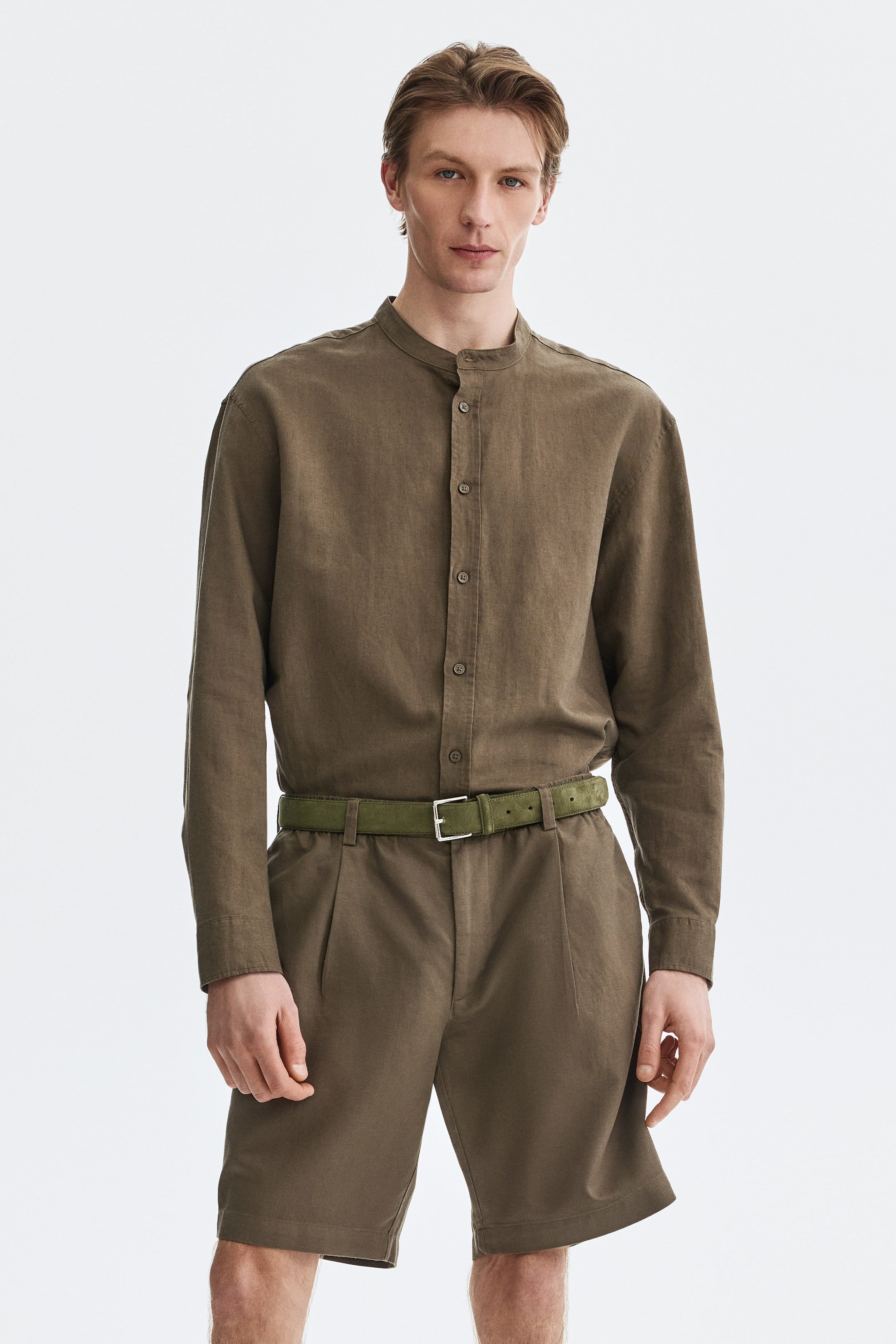 View larger image: "Presenting an olive green linen shirt with a band collar and button-down front, styled from the front view. It is paired with matching pleated shorts that feature an elasticated waist. The ensemble is completed with a green suede belt, accentuated by a silver-toned buckle, cinching the shorts perfectly."