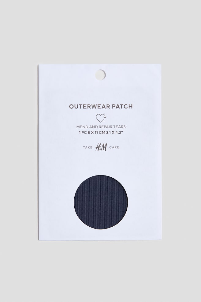 An outerwear patch package is shown from the front, with a dark navy blue, ribbed-textured fabric patch visible through a circular cutout in the lower half of a white cardboard sleeve. The upper portion of the sleeve displays printed text including "OUTERWEAR PATCH" and other product details. - 1