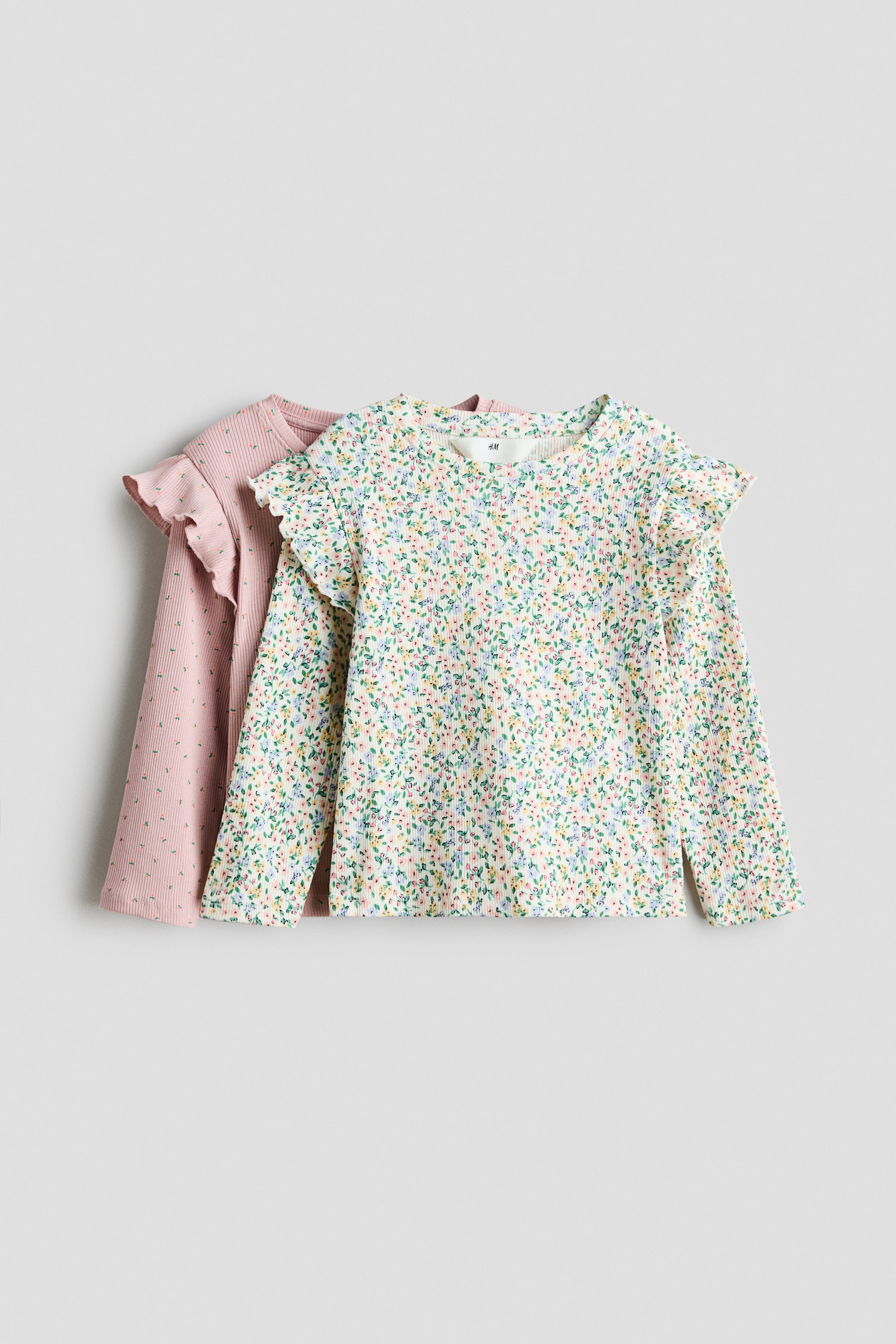 Ver imagen más grande: Dos blusas de manga larga acanalada se presentan de frente. La blusa delantera presenta una base blanca con un patrón floral multicolor y hombros con volantes, mientras que la blusa trasera parcialmente visible es rosa con un patrón de puntos sutil y hombros con volantes a juego.