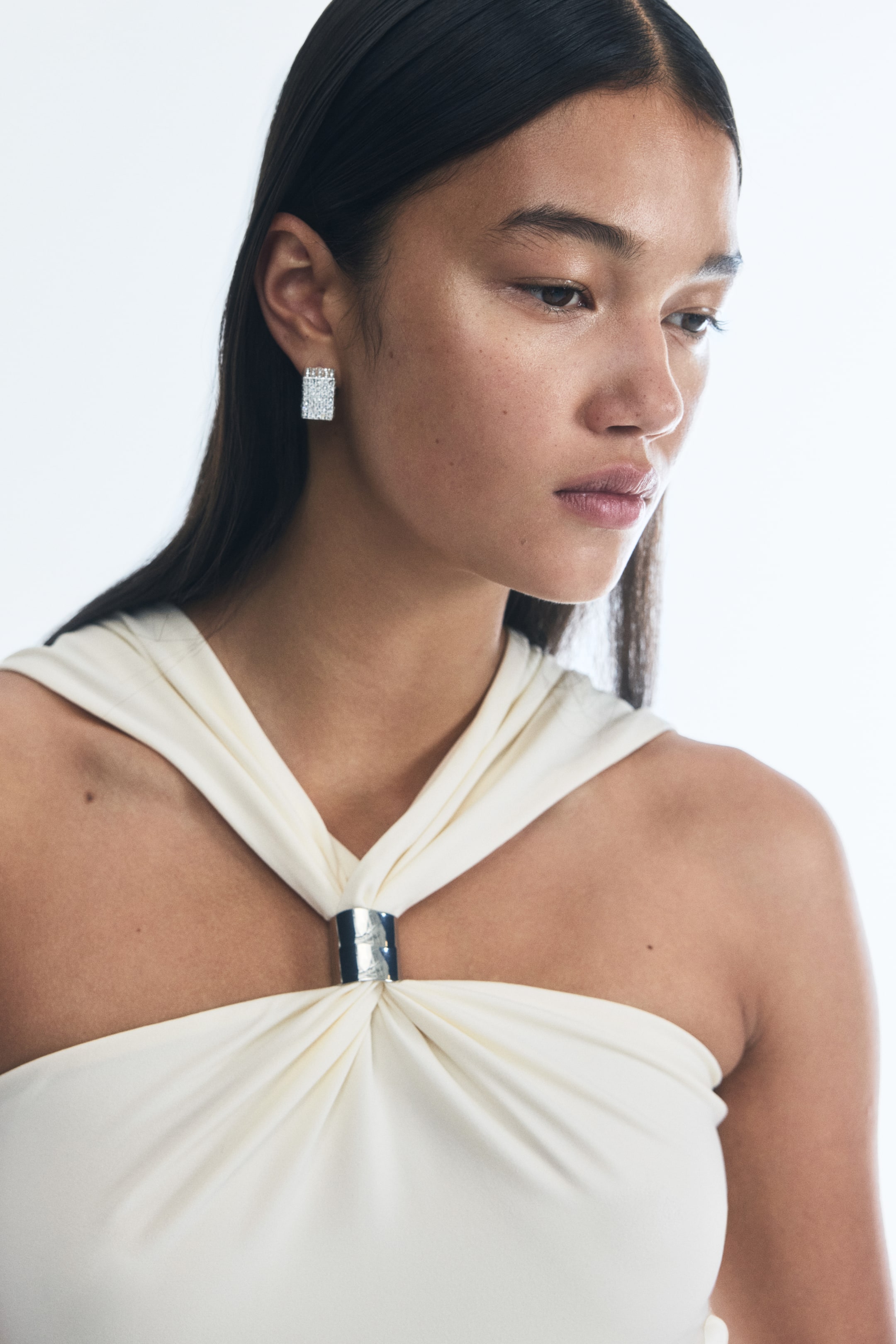 View larger image: A rectangular, pavé-set stud earring is worn on the ear, facing forward. A cream-colored garment is featuring a gathered neckline, where fabric is twisting and held by a rectangular metallic ring at the center of the chest.