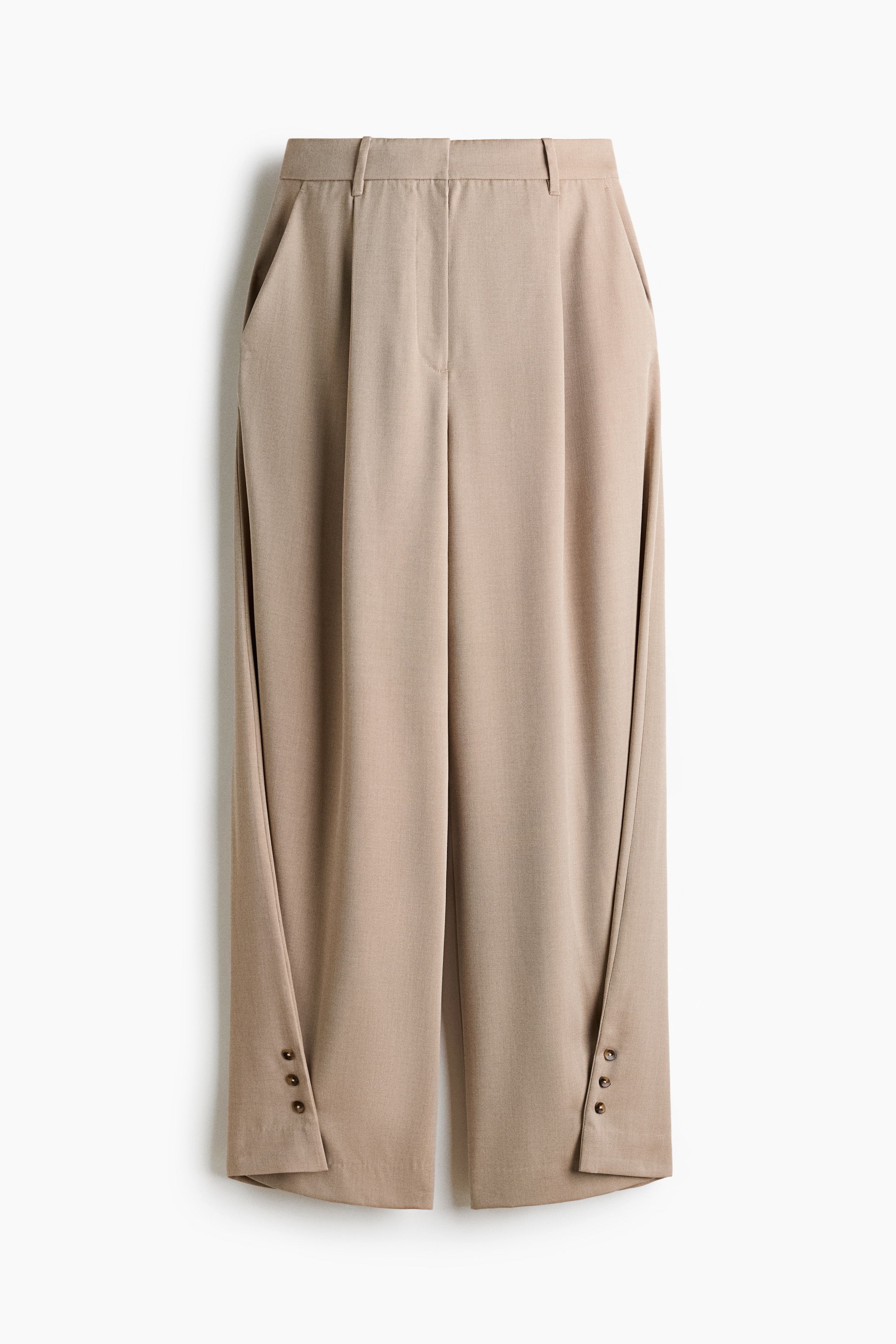 Pantalones tejidos color beige con cintura alta, pinzas delanteras y bolsillo lateral diagonal, usados por la modelo. Los pantalones anchos de pierna tipo barrel presentan un dobladillo cruzado con detalle de tres botones y se combinan con un suéter tejido color crema y una bolsa negra.