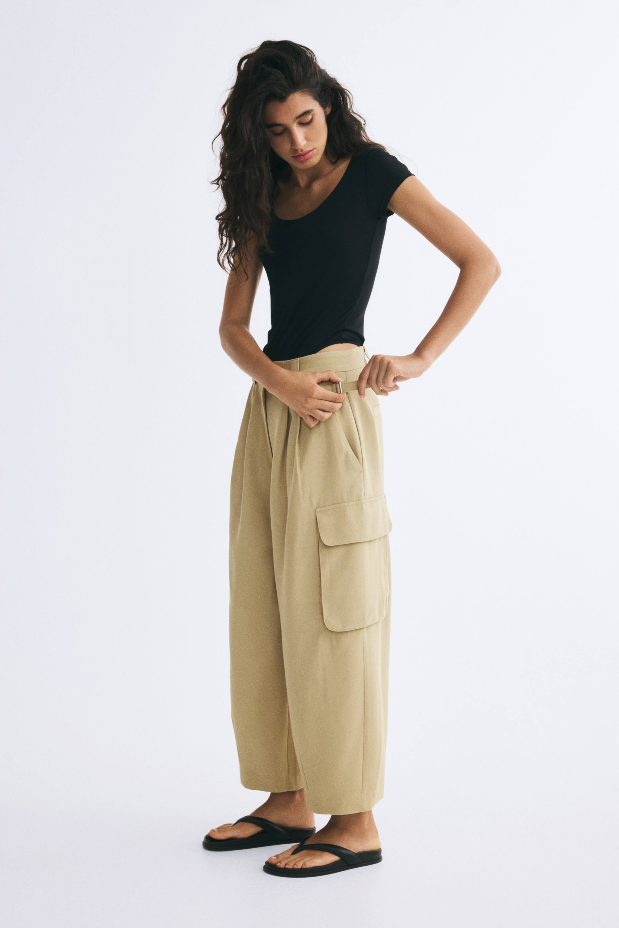 View larger image: Beige wide-leg cargo trousers with a side utility pocket and adjustable waist tabs are worn by the model, who is adjusting the side buckle strap with their hands. The trousers are styled with a fitted black short-sleeve top and black thong sandals.