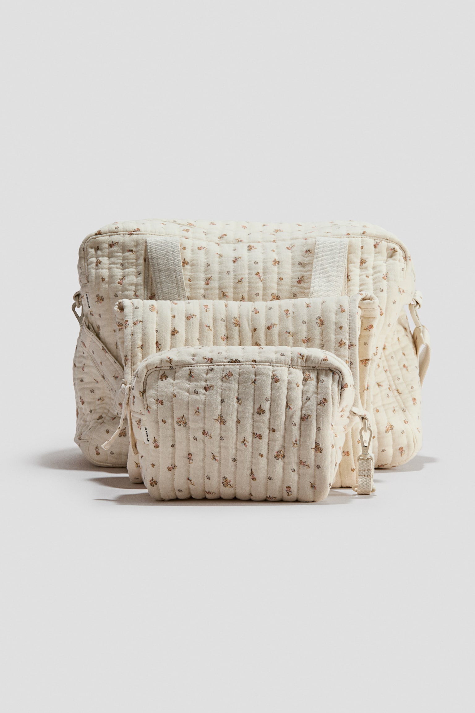 Ein dreiteiliges, gepolstertes Wickeltaschen-Set ist von vorne zu sehen und umfasst eine groÃe Wickeltasche, eine zusammengefaltete Wickelunterlage und eine kleinere Kulturtasche, die von vorne nach hinten angeordnet sind. Alle Teile sind aus cremefarbener, gesteppten Baumwoll-Musselin mit zartem, durchgehendem Blumenmuster gefertigt und verfügen über vertikale Steppnähte, ReiÃverschlüsse und Riemenbefestigungen. - 1