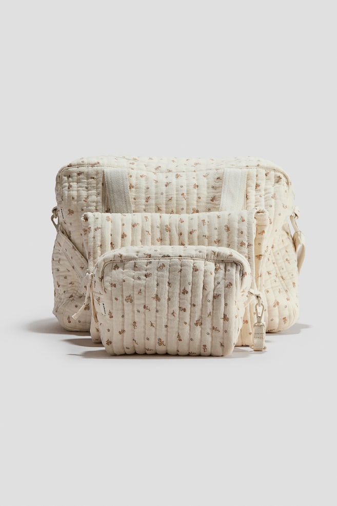 A three-piece padded nappy bag set is shown from the front, featuring a large nappy bag, a folded changing mat, and a smaller toiletry bag arranged front-to-back. All pieces are crafted from cream-colored quilted cotton muslin with a delicate, all-over floral print and feature vertical quilting lines, zippers, and strap attachments. - 1
