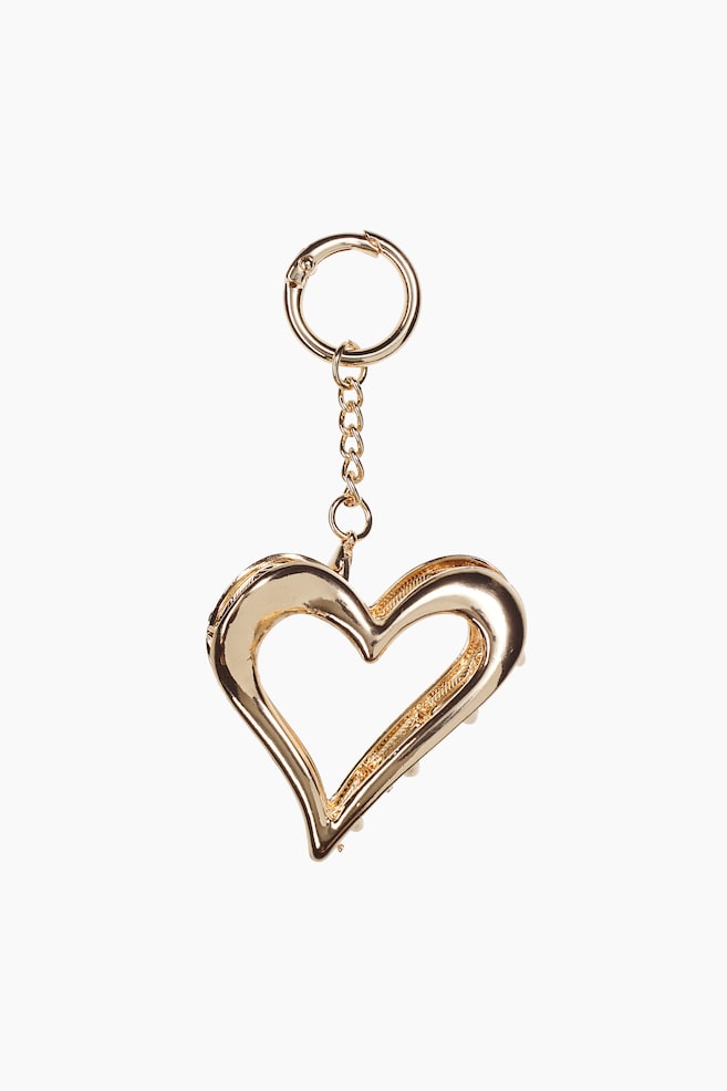 Ein goldfarbenes Schlüsselanhänger-Accessoire hängt vertikal und zeigt eine hohle, herzförmige Krallenklammer, die an einer kurzen Gliederkette und einem runden Federring befestigt ist. Die polierte Metallklammer ist nach vorne ausgerichtet und präsentiert ihre glatte, reflektierende Oberfläche sowie die dezent sichtbaren Krallenzähne entlang der Innenkante. - 1