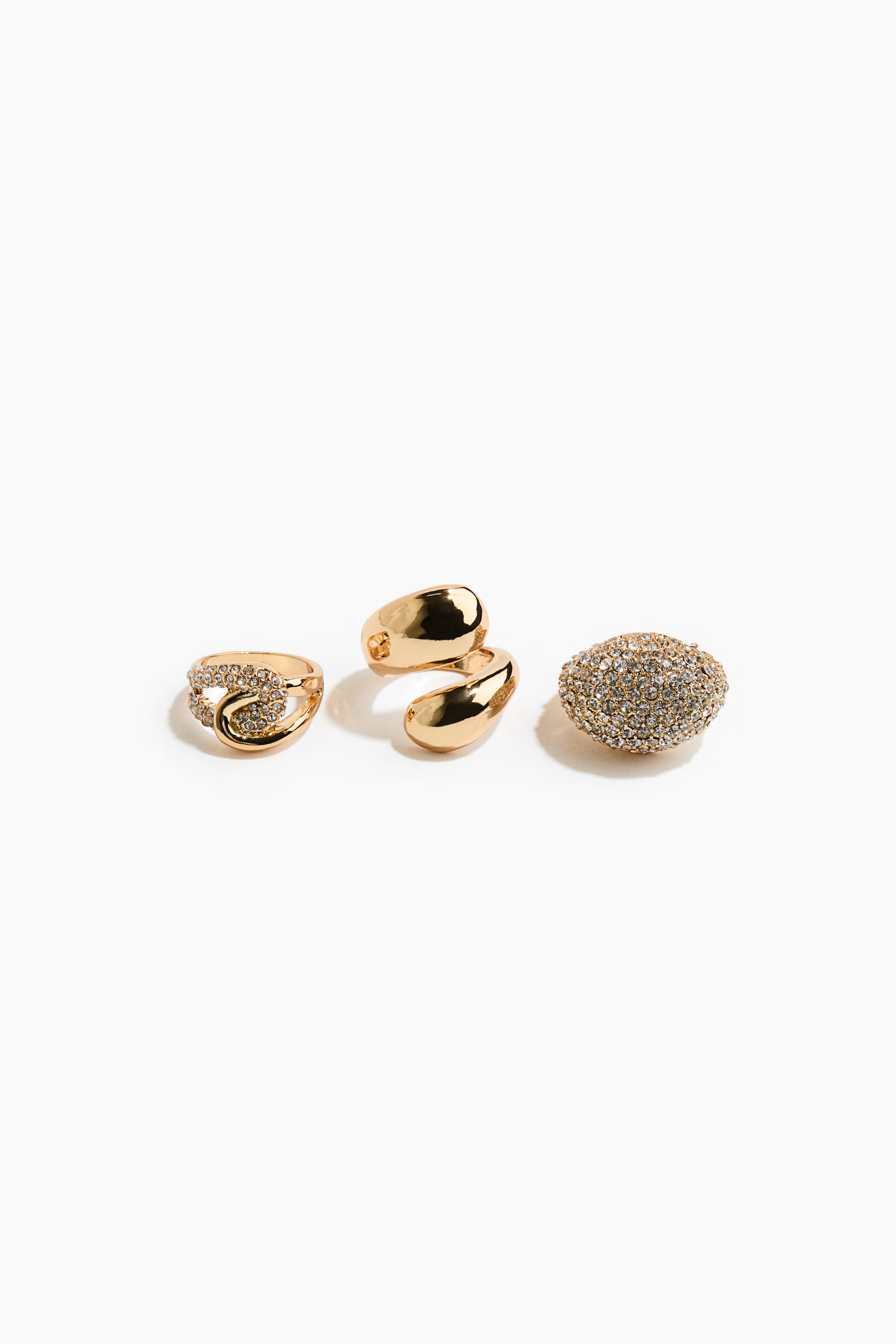 Visualizza l'immagine ingrandita: Tre anelli color oro sono disposti fianco a fianco in una vista frontale, mettendo in evidenza design distinti. Il set comprende un anello con fascia intrecciata impreziosita da strass, un anello bypass lucido con estremità arrotondate e un ampio anello cocktail a cupola, ricoperto di cristalli trasparenti pavé.