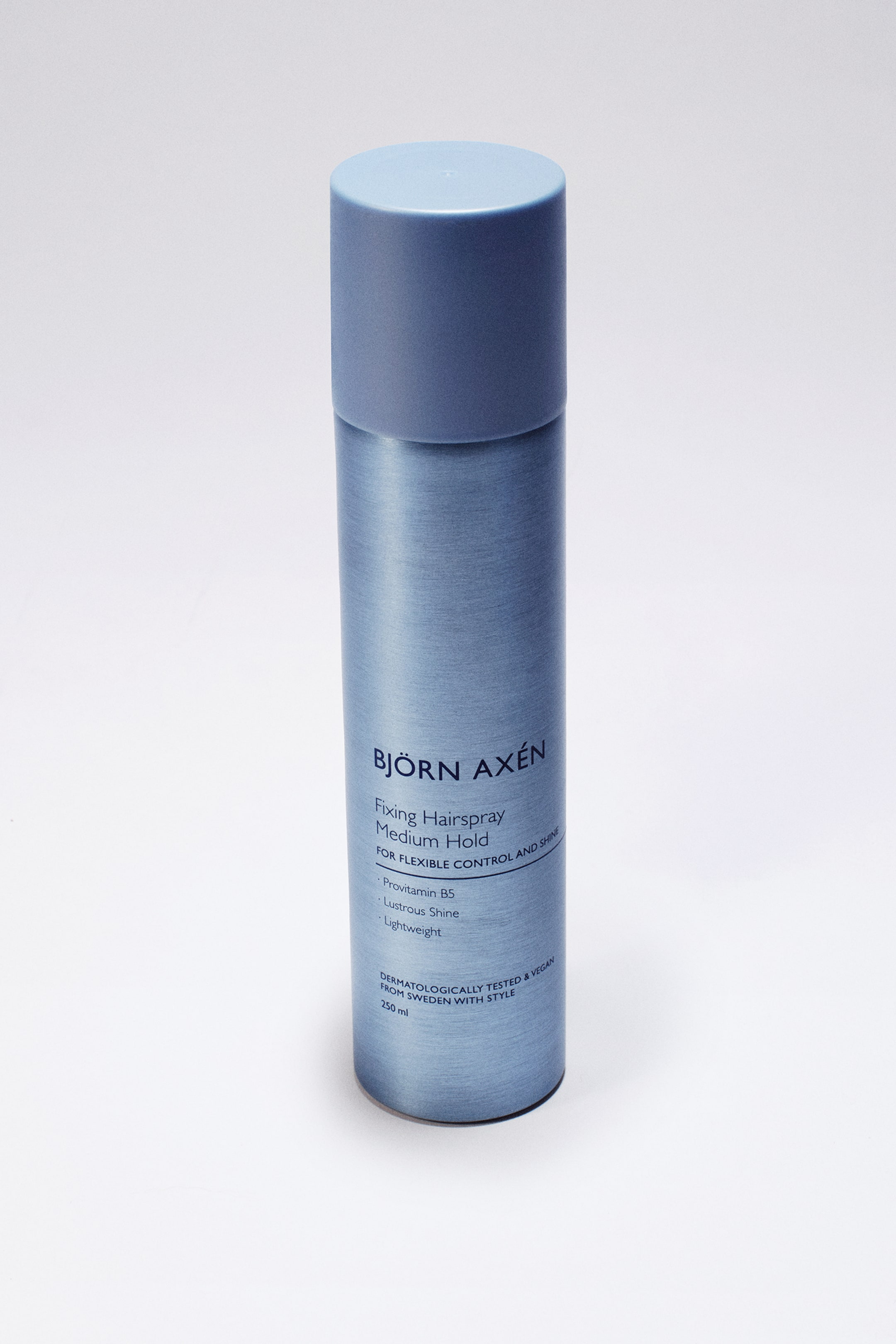 Vis større bilde: En boks Björn Axén Fixing Hairspray står oppreist, med en sylindrisk kropp i metallisk blågrå med børstet finish og en matchende solid matt blå kork. Den vendte etiketten har mørkeblå tekst som viser at dette er en formel med «Medium Hold» og oppgir nøkkelfunksjoner som «Provitamin B5» og «Lustrous Shine».