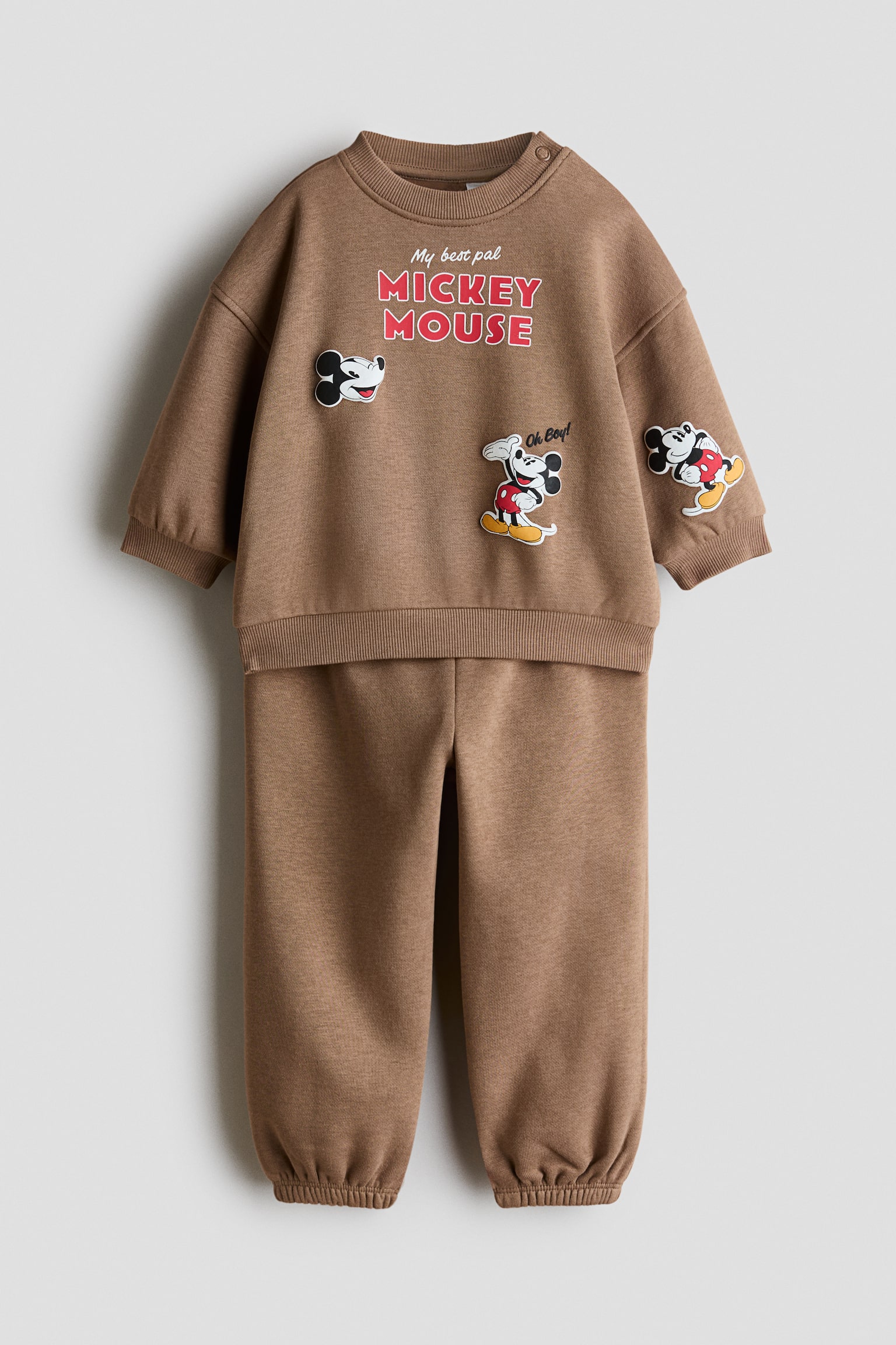 A brown 2-piece printed sweat set is shown from the front, featuring a long-sleeved sweatshirt and matching sweatpants. The sweatshirt has a ribbed crew neck with a shoulder snap button, ribbed cuffs, and Mickey Mouse graphic prints, while the coordinating sweatpants feature gathered elastic cuffs at the ankles.