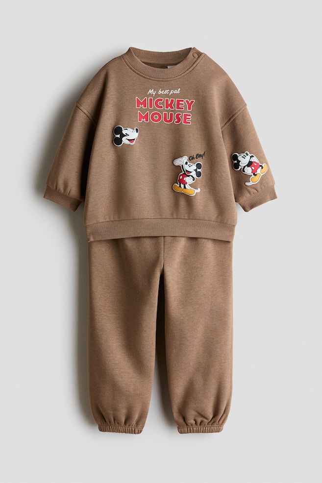 Ett brunt 2-delat set i sweat-tyg med print visas framifrån och består av en långärmad sweatshirt och matchande sweatpants. Sweatshirten har en ribbad rund hals med tryckknapp vid axeln, ribbade muddar och Mickey Mouse-grafiska tryck, medan de matchande sweatpantsen har resårmuddar vid anklarna. - 1
