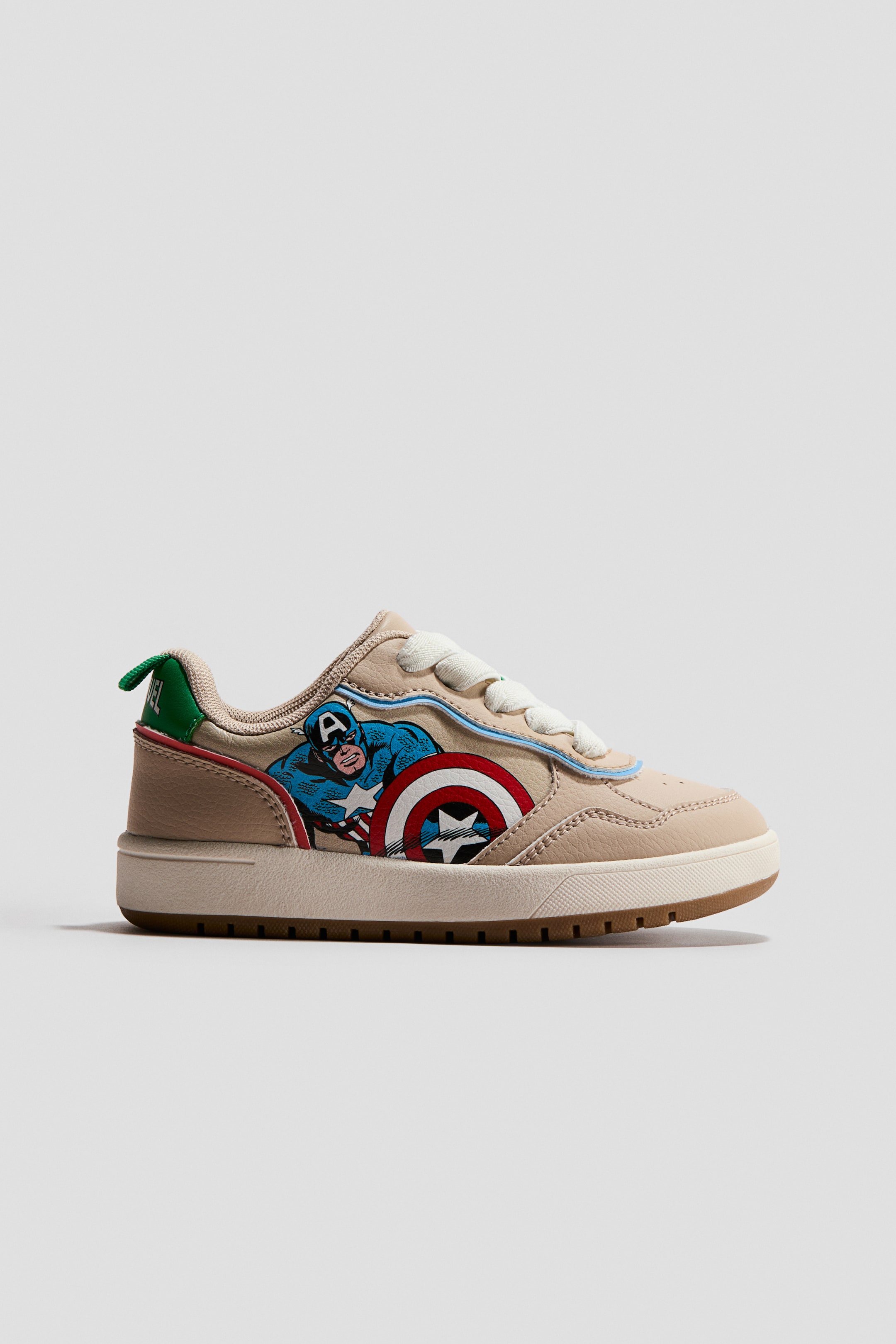 View larger image: A beige low-top sneaker is facing FRONT, featuring a Captain America graphic on its outer side. White laces, light blue piping, and red piping are visible, along with a green heel tab displaying "MARVEL" text. A cream midsole with a brown textured outsole is present.