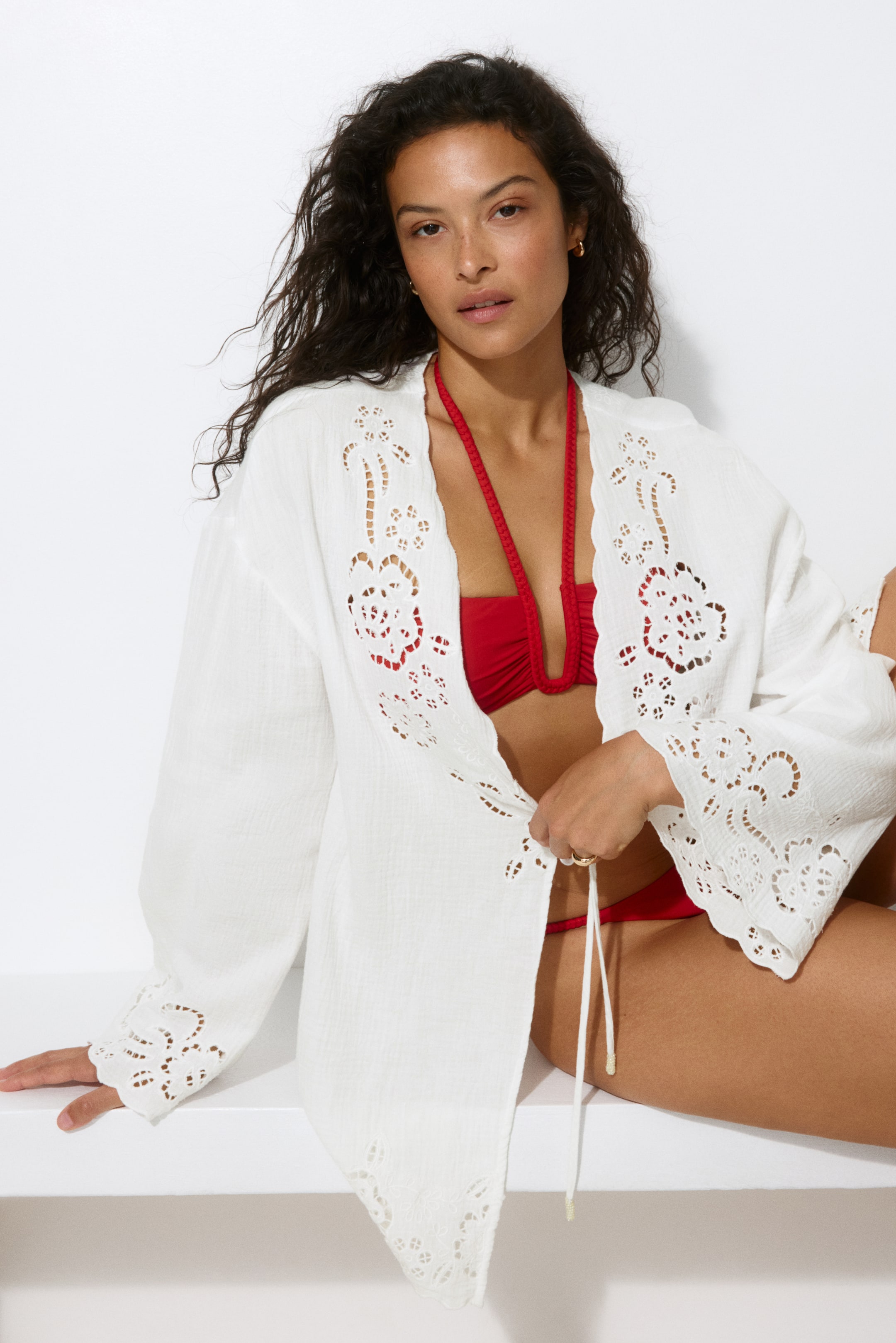 Visualizza immagine più grande: La modella indossa un corto caftano da spiaggia a vestibilità ampia in morbido cotone mussola con broderie anglaise sopra un bikini rosso. Presenta una profonda scollatura a V, un'apertura frontale con lacci stretti e maniche lunghe, con la modella che tiene uno dei lacci anteriori.