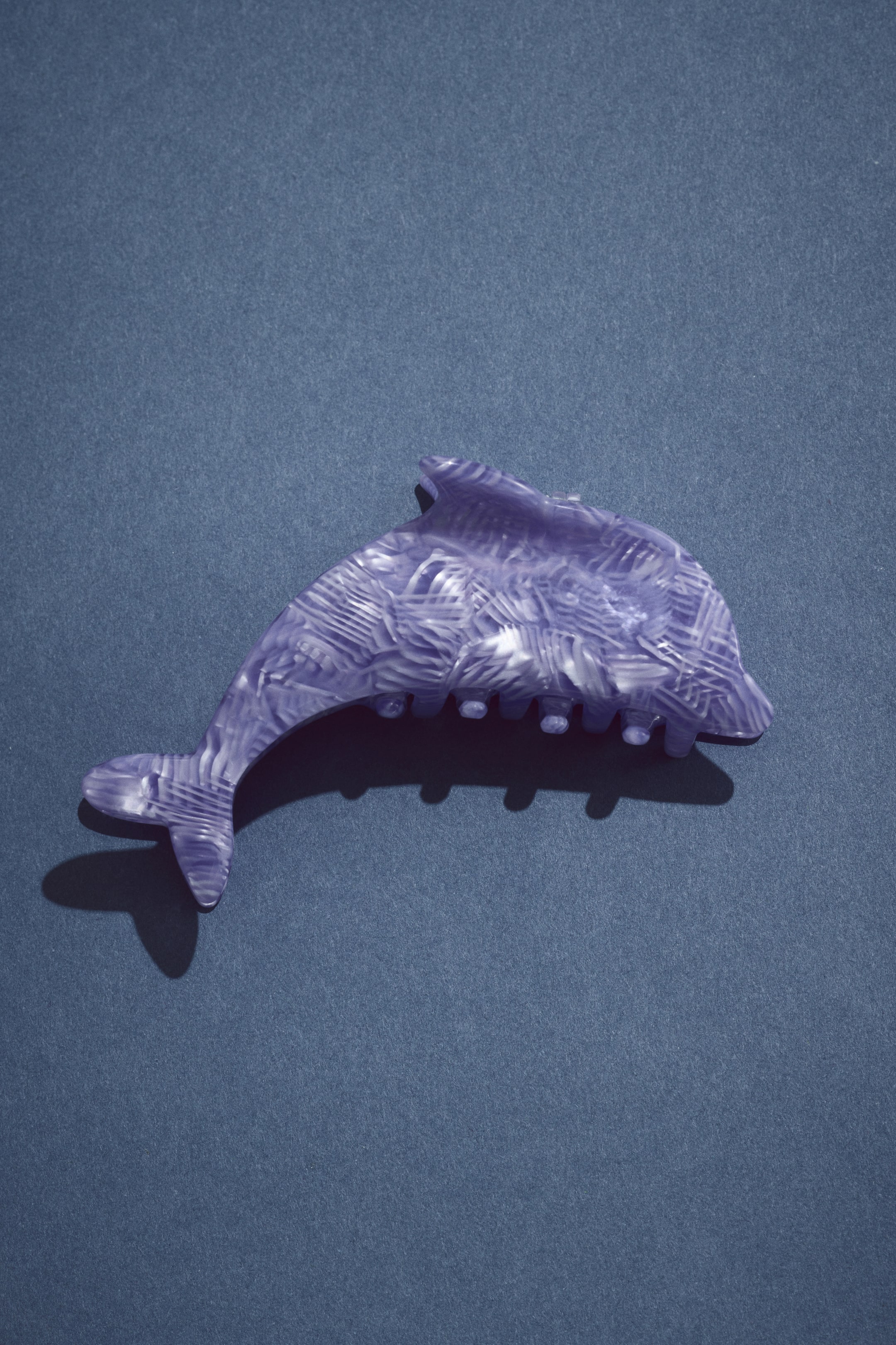 View larger image: A dolphin-shaped hair claw clip is facing left, featuring a marbled lilac purple and white pattern. Its clamping teeth are visible along the underside.