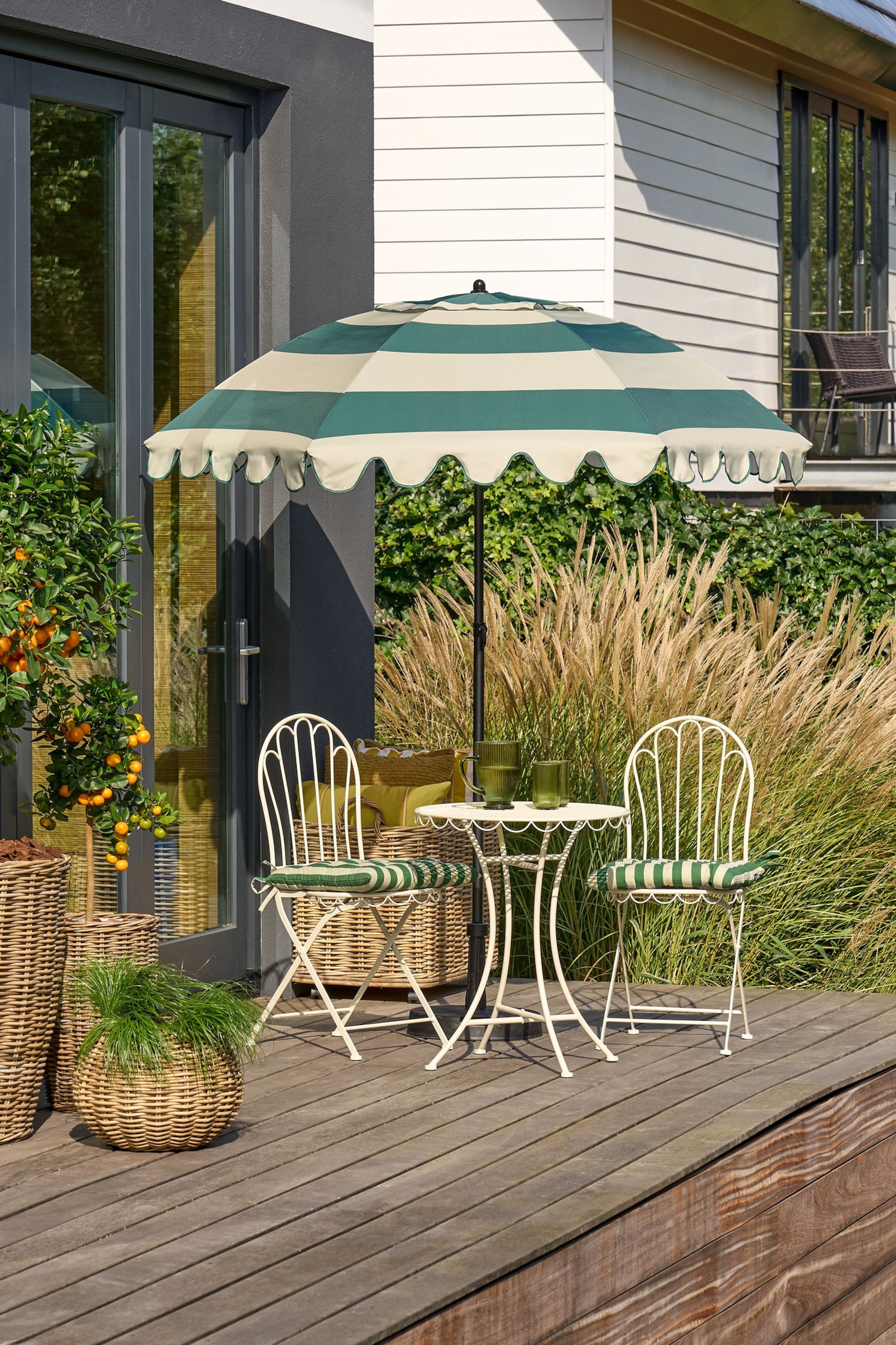Größeres Bild anzeigen: Ein Outdoor-Patio-Set präsentiert einen grün- und cremegestreiften Sonnenschirm mit gewelltem Rand, einen kleinen runden Tisch aus cremefarbenem Metall und zwei passende Stühle aus cremefarbenem Metall. Die Stühle sind mit grün- und cremegestreiften Kissen ausgestattet, während zwei grüne gerippte Gläser auf dem Tisch stehen.
