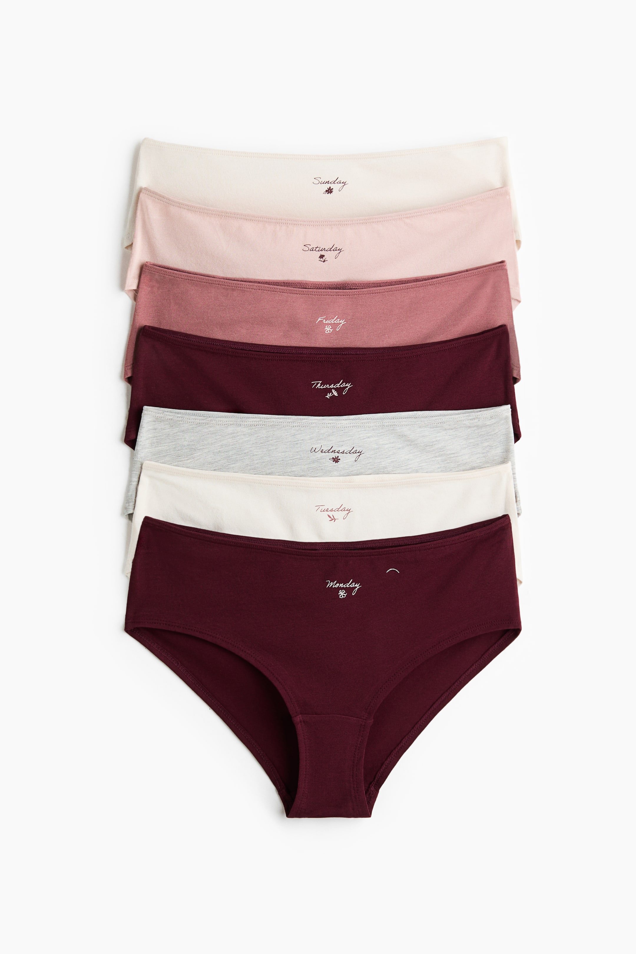 Visualizza immagine più grande: Sette slip in jersey sono impilati, rivolti verso il fronte e mostrano una gamma di colori tenui tra cui bordeaux, bianco sporco, grigio chiaro, rosa polvere e rosa chiaro. Ogni slip a vita media è ricamato con un giorno della settimana e un piccolo emblema floreale sulla parte anteriore della cintura, con il tassello foderato dello slip inferiore chiaramente visibile.