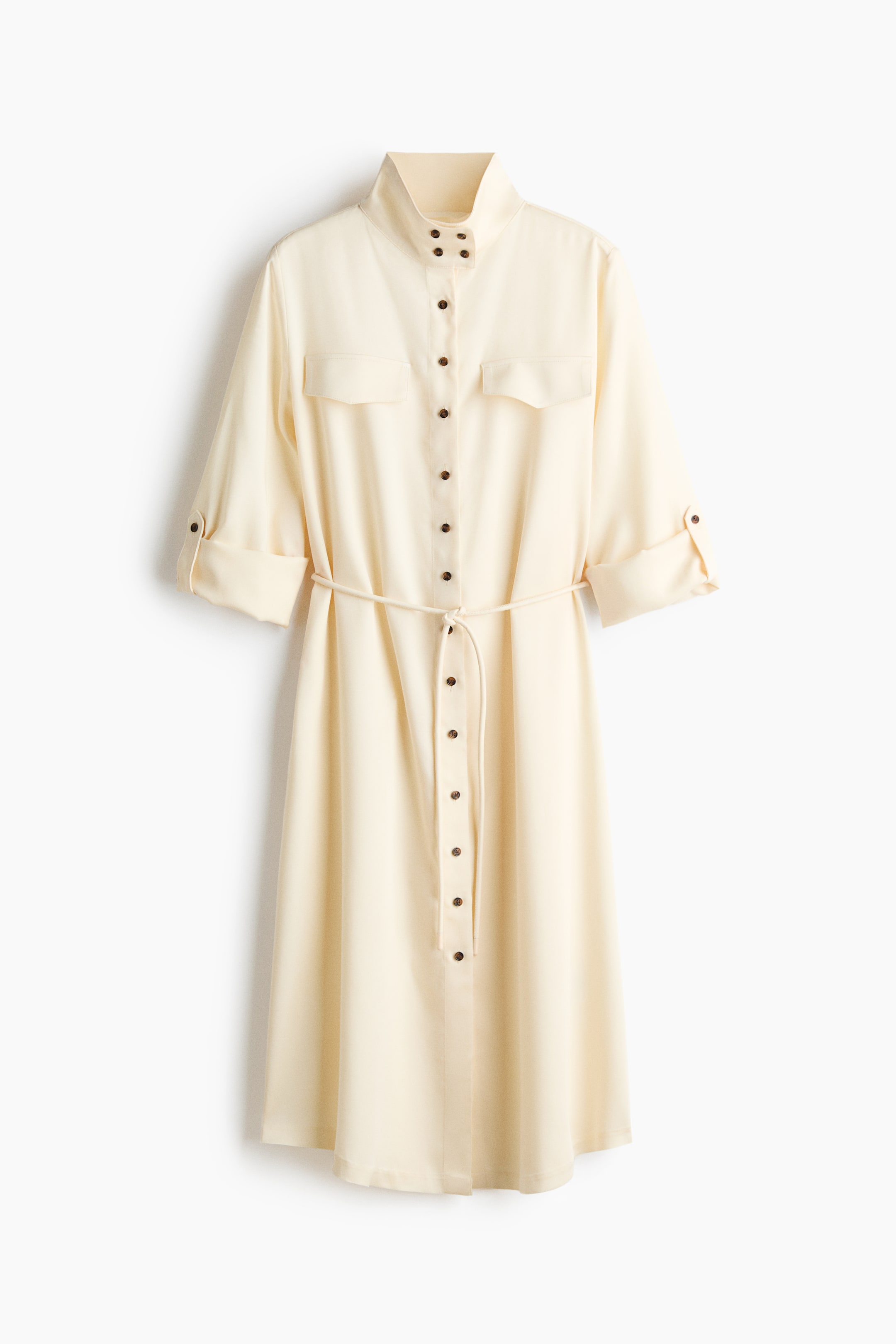 View larger image: A cream midi shirt dress is displayed frontally, featuring a band collar and a full button-front placket with dark buttons. It is designed with two chest flap pockets, three-quarter length sleeves rolled up and secured with tabs, and a self-fabric tie belt cinching the waist.