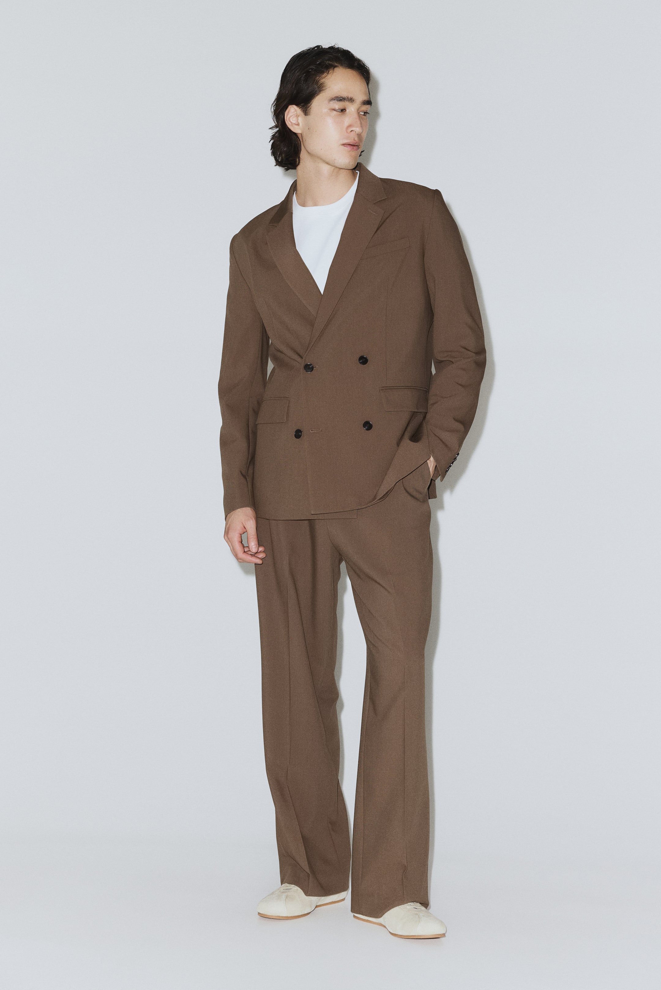 View larger image: A brown double-breasted suit features a blazer with peak lapels, two rows of dark buttons and flap pockets, worn over a white T-shirt. The matching trousers have a relaxed, straight-leg fit, completing the look with light-coloured flats.