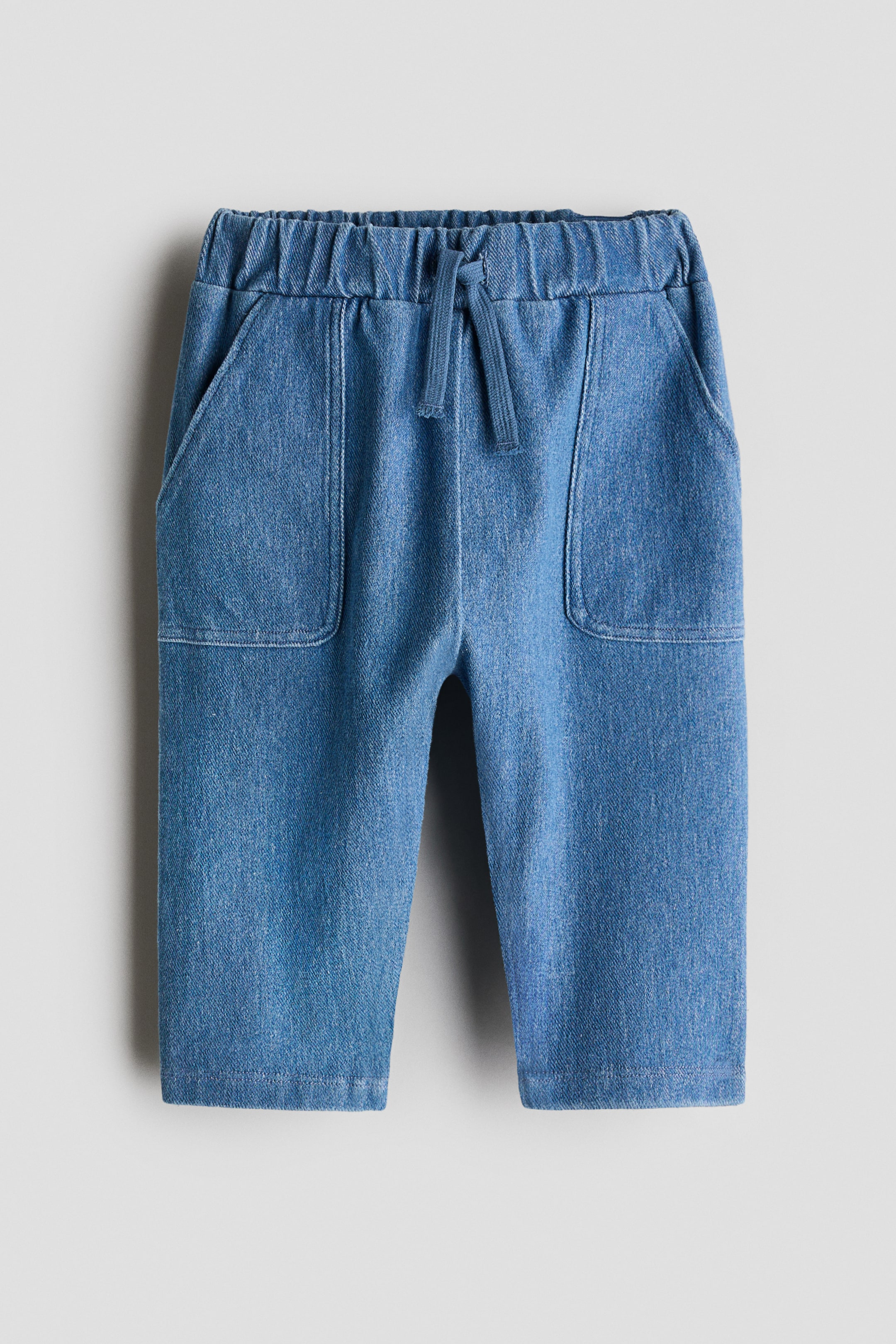 View larger image: Front-facing denim-look trousers feature an elasticated, gathered waist with a decorative drawstring, two vertical seams running down the front, and two large patch pockets on the front.