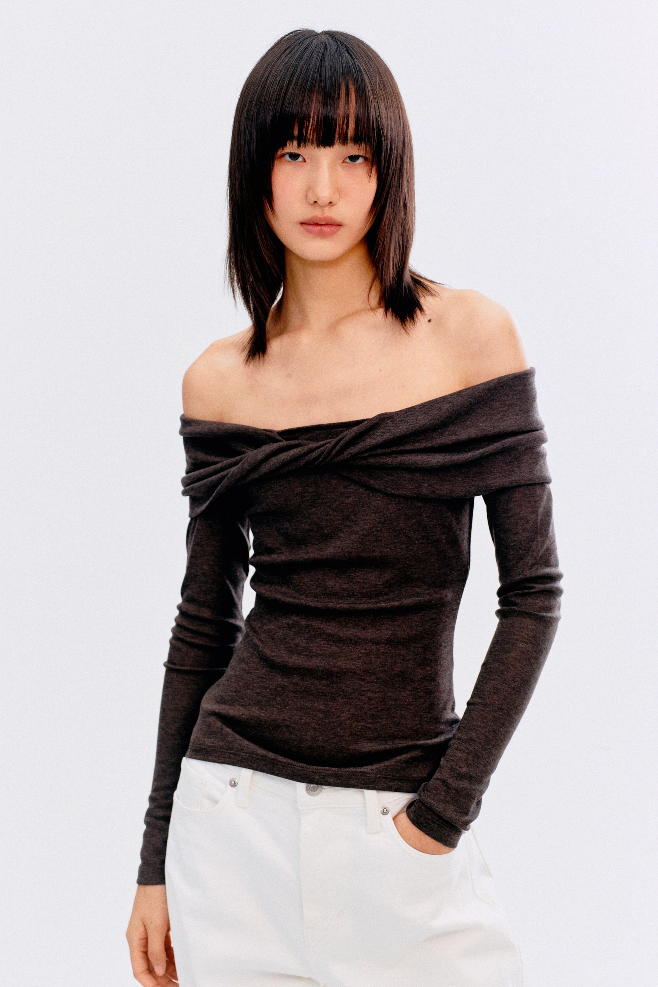拡大画像を表示: ダークグレーのヘザードニットオフショルダートップは、ツイストネックラインのディテールと長袖が特徴で、前面から見るとフィットしたシルエットを演出します。