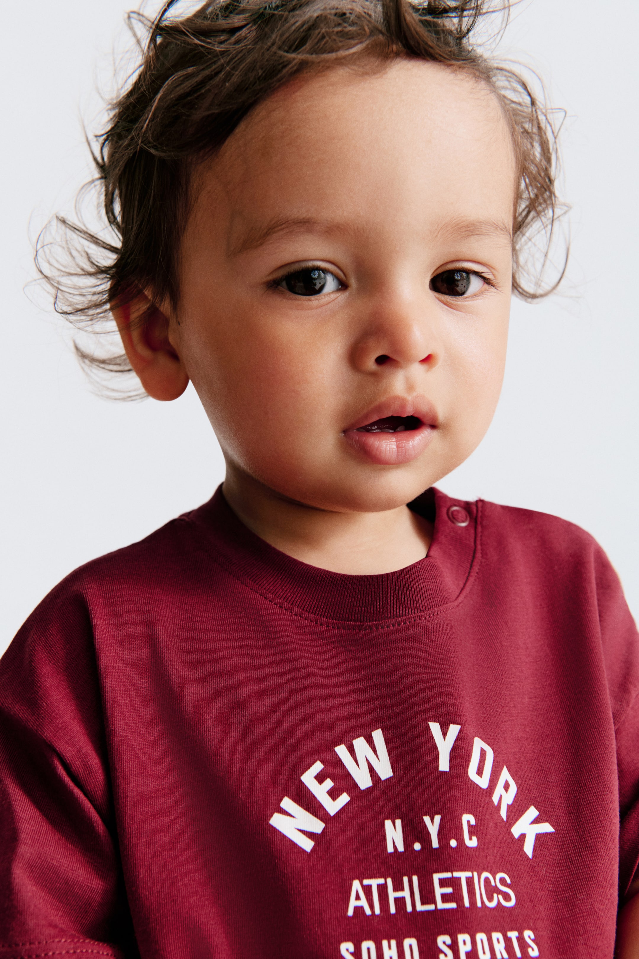 View larger image: A maroon t-shirt features a white text graphic on the front, reading 'NEW YORK' in an arch, followed by 'N.Y.C', 'ATHLETICS', and 'SOHO SPORTS'. It also showcases a ribbed crew neck and two snap buttons on the left shoulder.