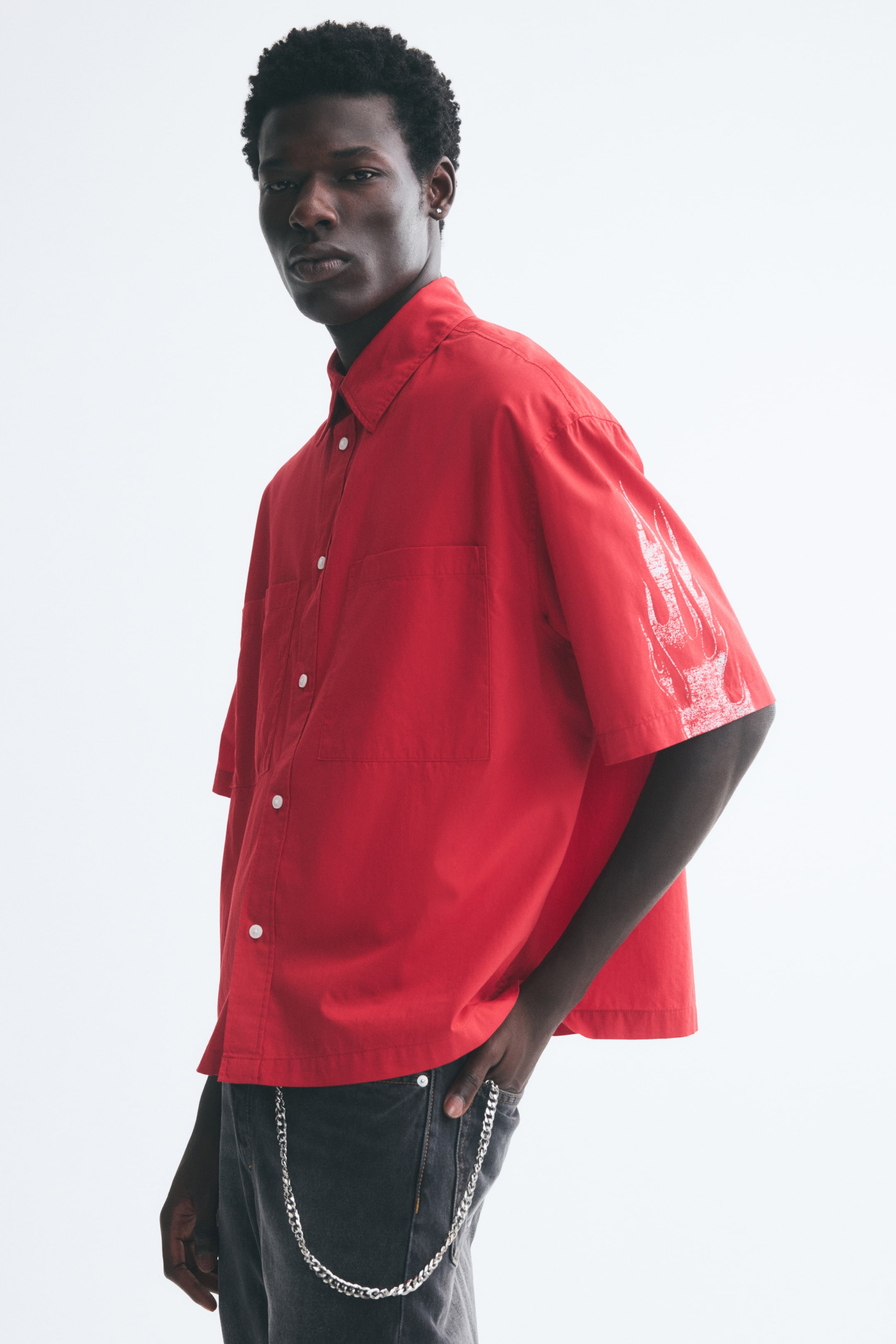View larger image: A red, short-sleeve button-down shirt in a relaxed fit is being worn by the model, featuring a classic collar, a patch chest pocket, and a white printed flame graphic on the sleeve. The shirt is styled untucked over gray jeans with a silver metal chain accessory hanging from the waistband.