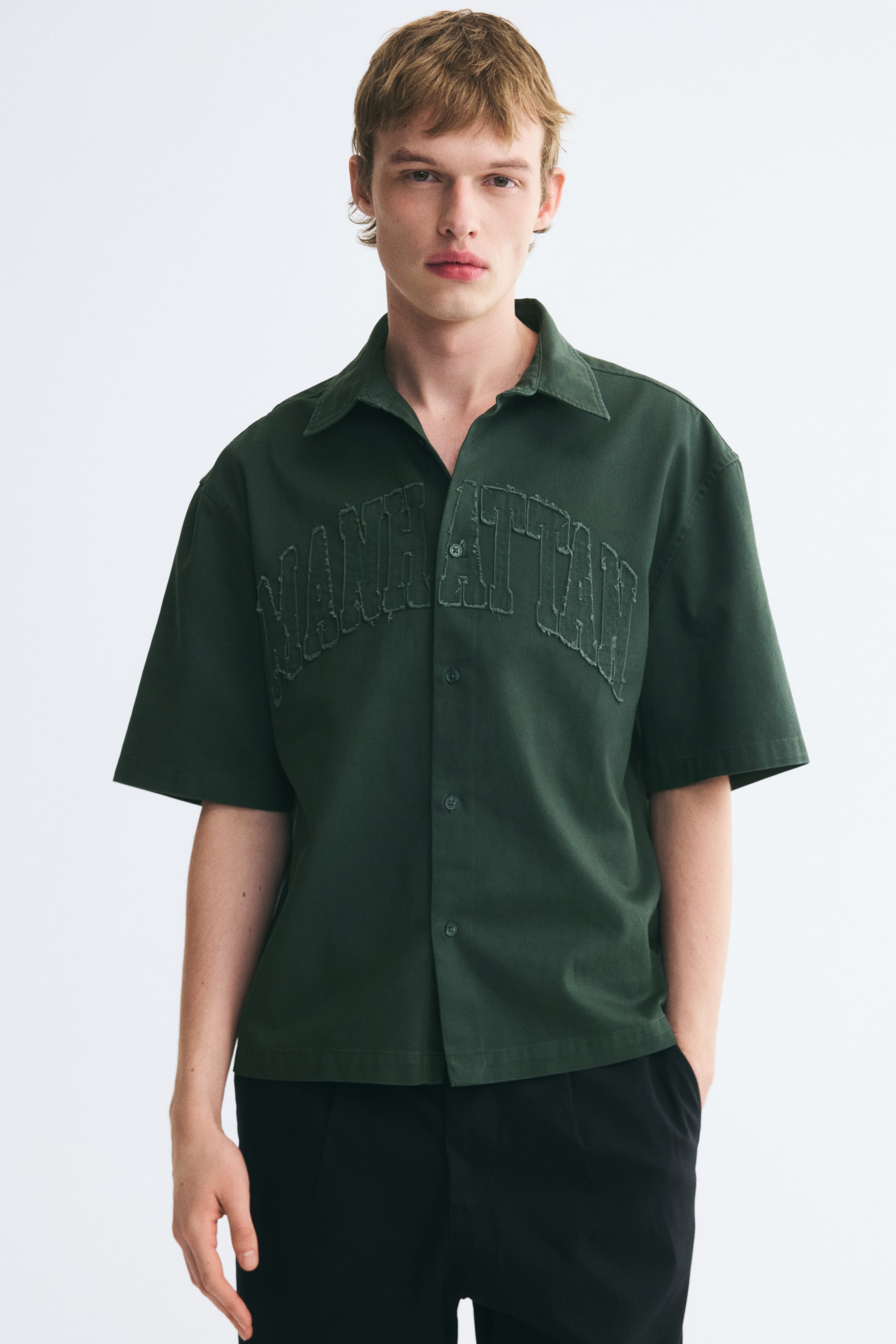 View larger image: The model is wearing a dark green, short-sleeved button-up shirt with a distressed applique spelling "MANHATTAN" across the chest, paired with black trousers.