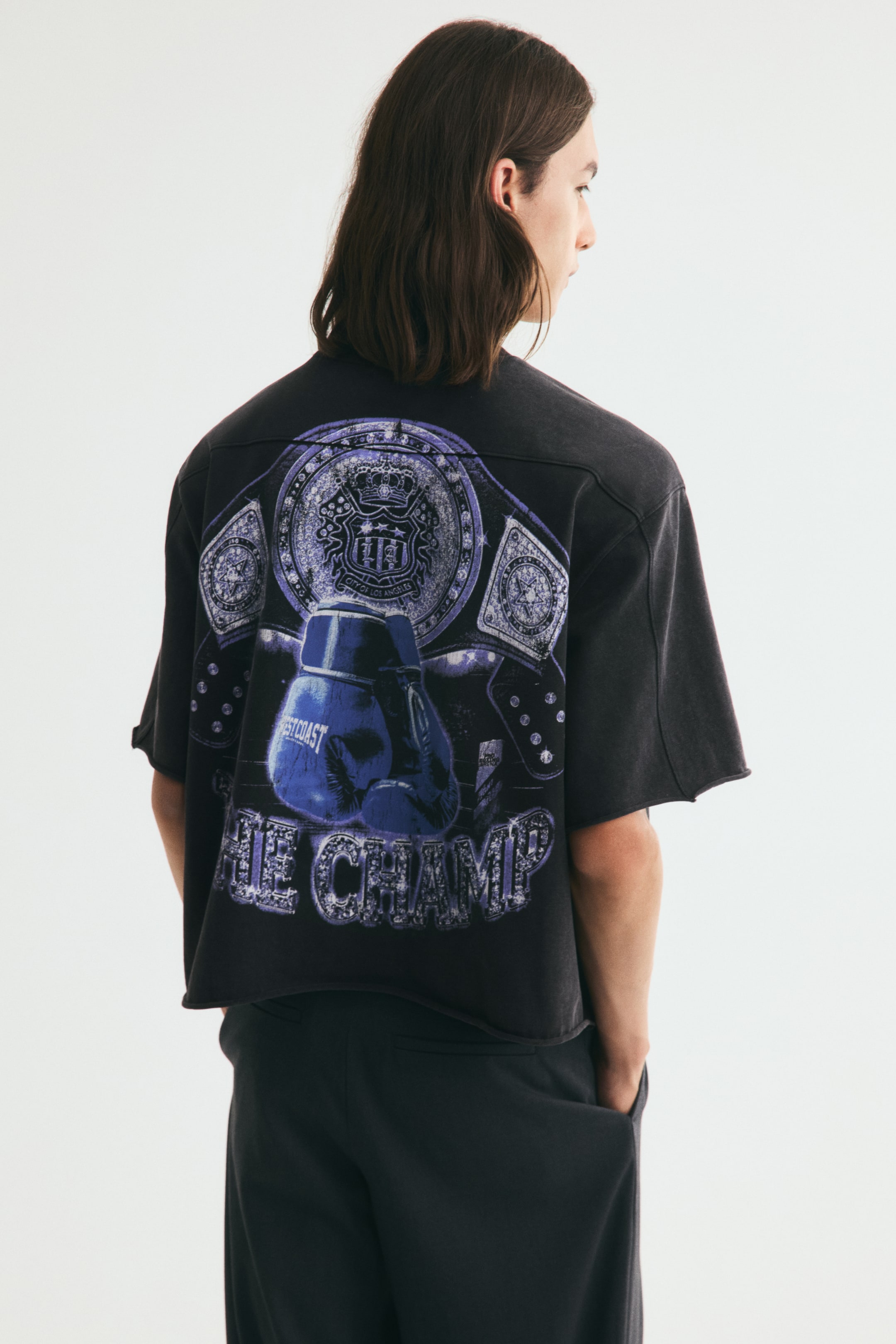 View larger image: A short, boxy black t-shirt with an oversized fit is being worn by the model, showcasing a large purple and blue graphic on the back featuring boxing gloves, a championship belt, and the text "THE CHAMP". The heavyweight jersey tee features dropped shoulders, wide sleeves, and raw, rolled edges at the cuffs and hem.