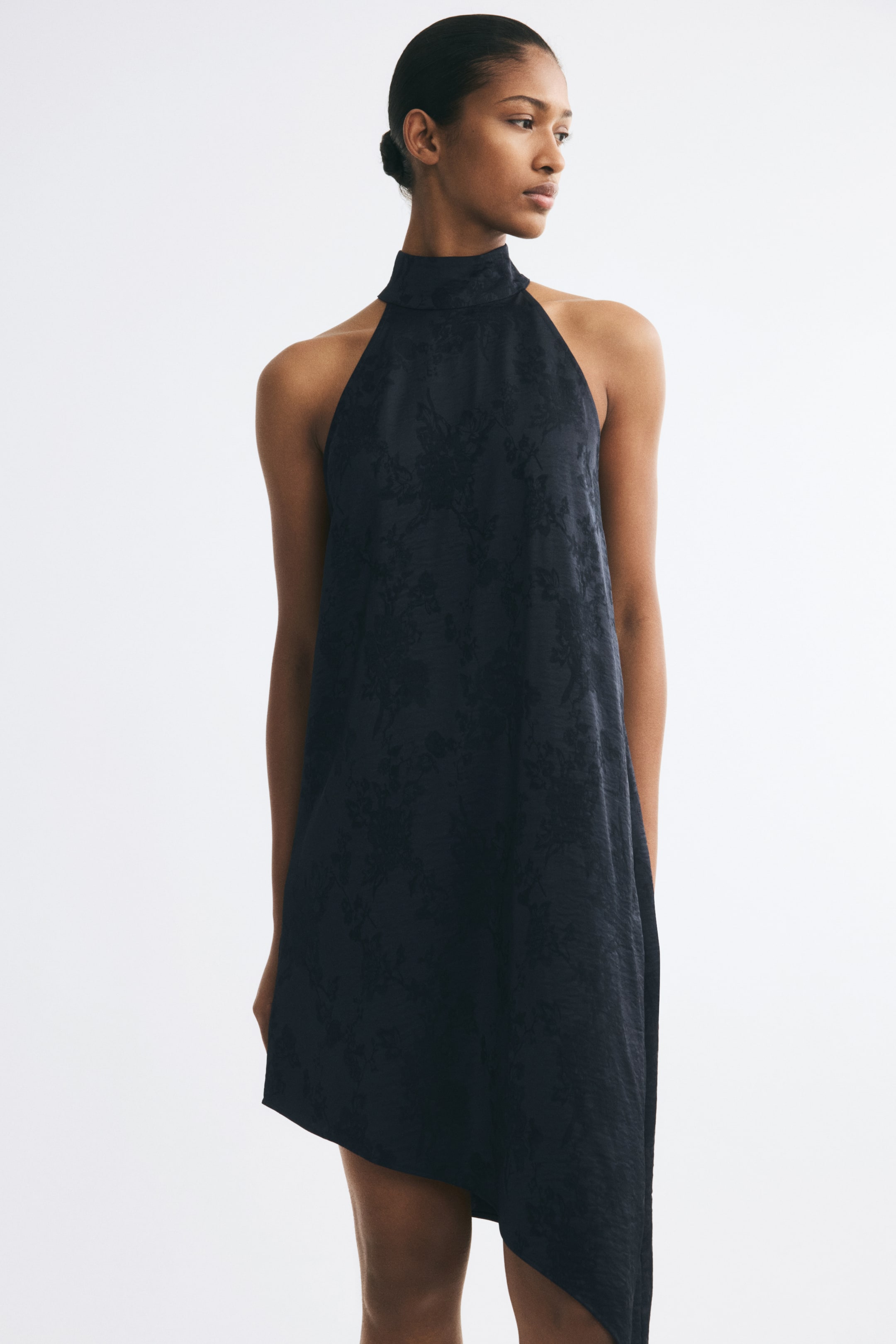 View larger image: The model is wearing a black sleeveless dress in a textured floral jacquard weave. The dress features a high stand collar with a halter-style neckline and a relaxed silhouette that ends in an asymmetrical hem.