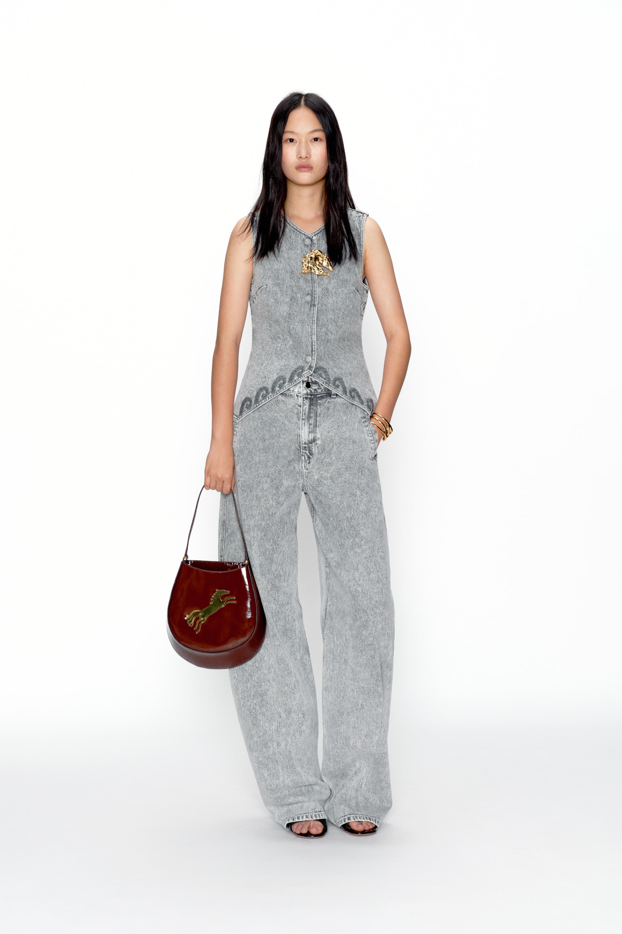 View larger image: A washed grey denim sleeveless vest paired with matching wide-leg trousers, featuring a V-neckline, button-front closure, and a pointed hem with dark grey embroidered detailing along the waist. Styled with a dark brown glossy half-moon handbag showcasing a gold leaping horse emblem.