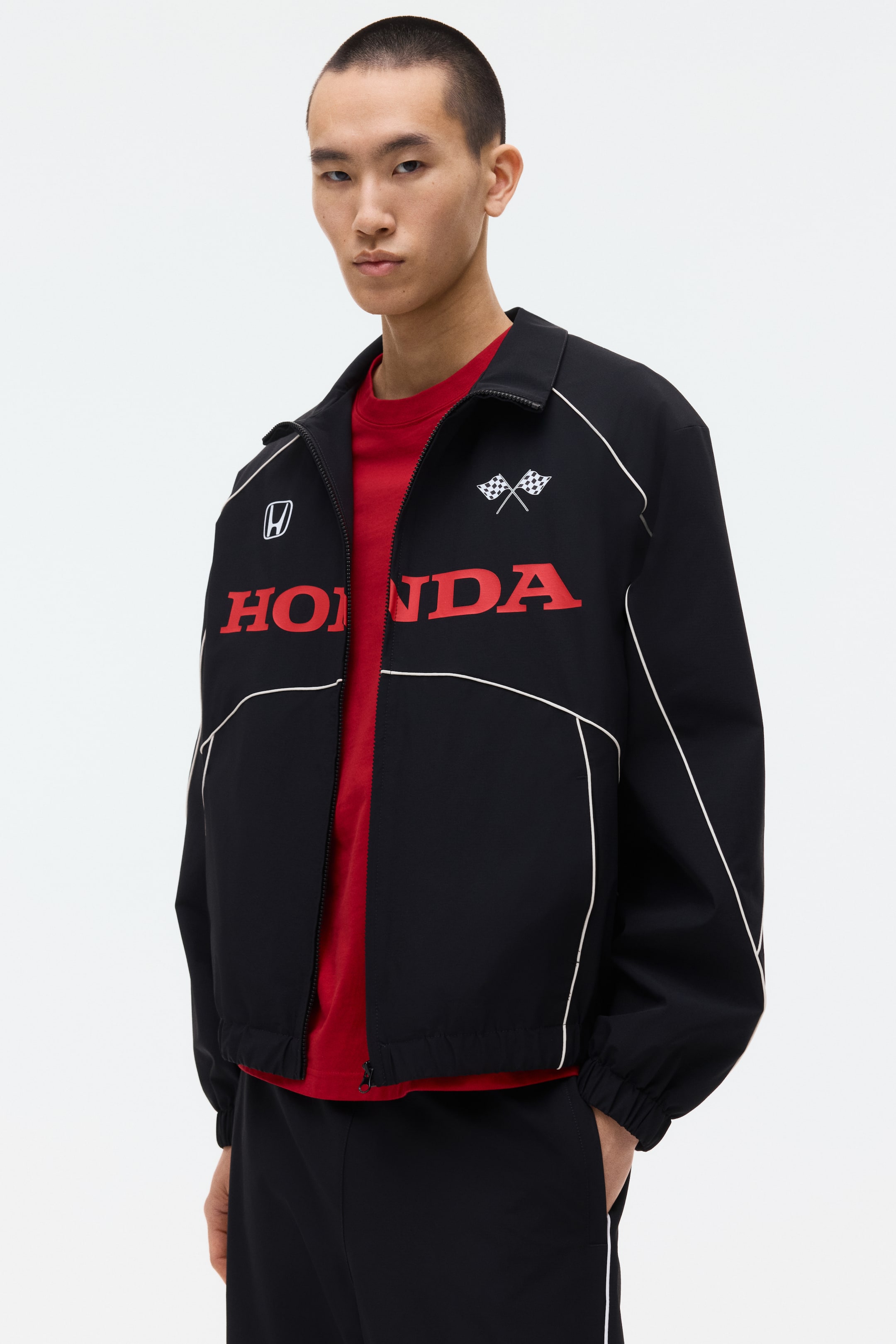 View larger image: A black racing jacket featuring a full-zip closure and a pointed collar. Red 'HONDA' lettering is displayed across the chest, with a white 'H' logo on the left and crossed checkered flags on the right, all outlined by white piping. The jacket, with elasticated cuffs and hem, is worn open over a red t-shirt.