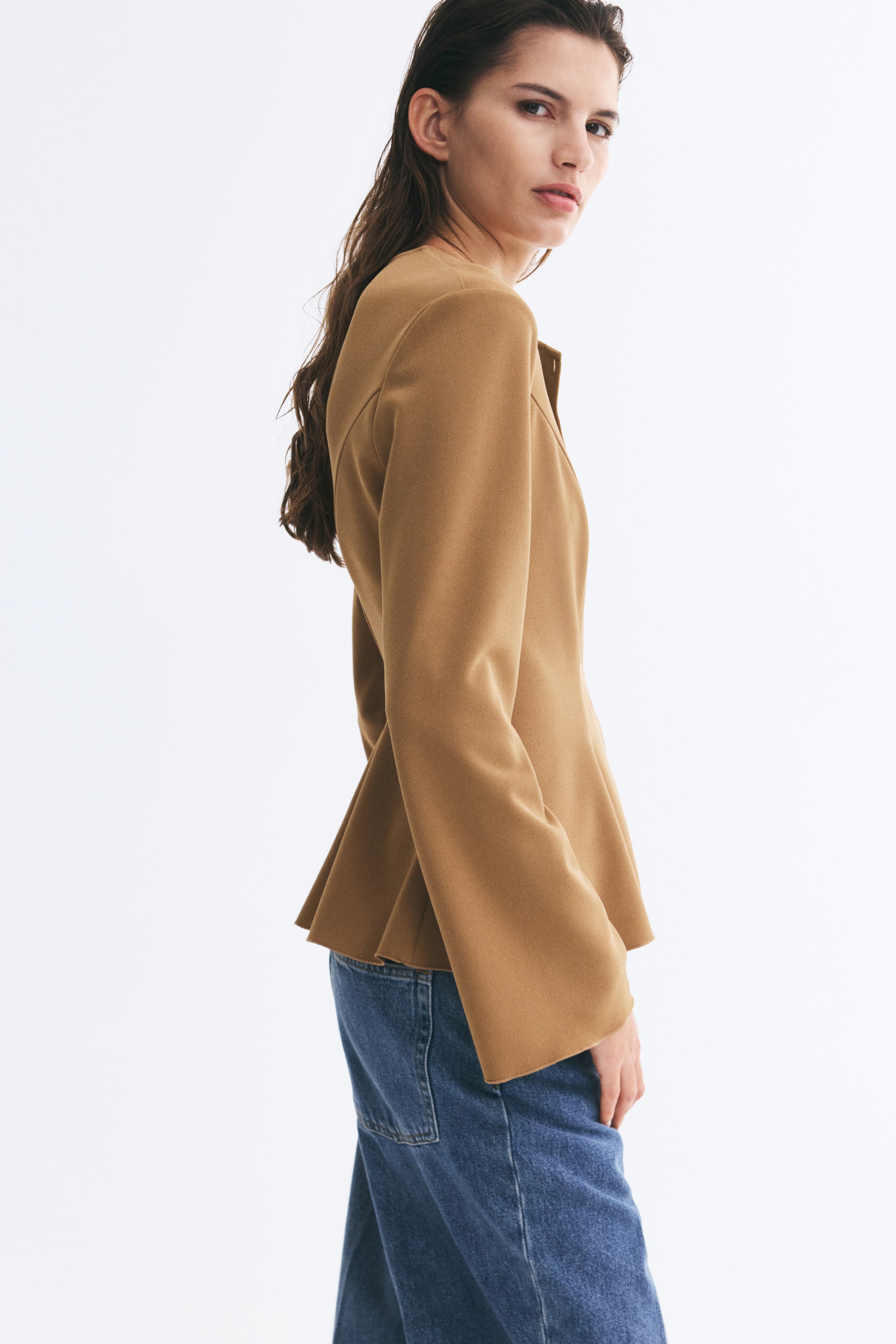 View larger image: A camel-coloured jacket is worn by the model, featuring a collarless neckline, flared long sleeves, and a structured peplum hem. The jacket is styled with blue jeans, showcasing its tailored side profile.