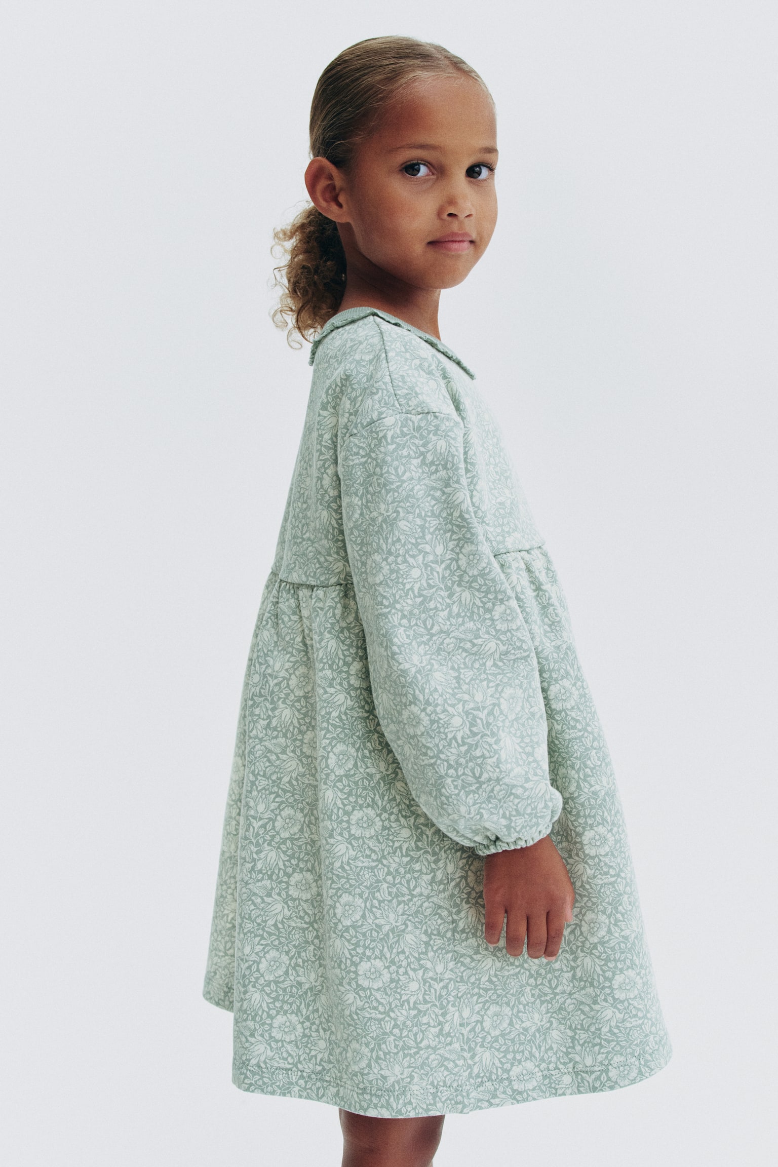 A light green long-sleeved dress is shown from the front, featuring an all-over white floral print. The dress is designed with a round, rib-trimmed neckline displaying a delicate lace trim, dropped shoulders, voluminous sleeves with elasticated cuffs, and a gathered waist flowing into a flared skirt. - 1