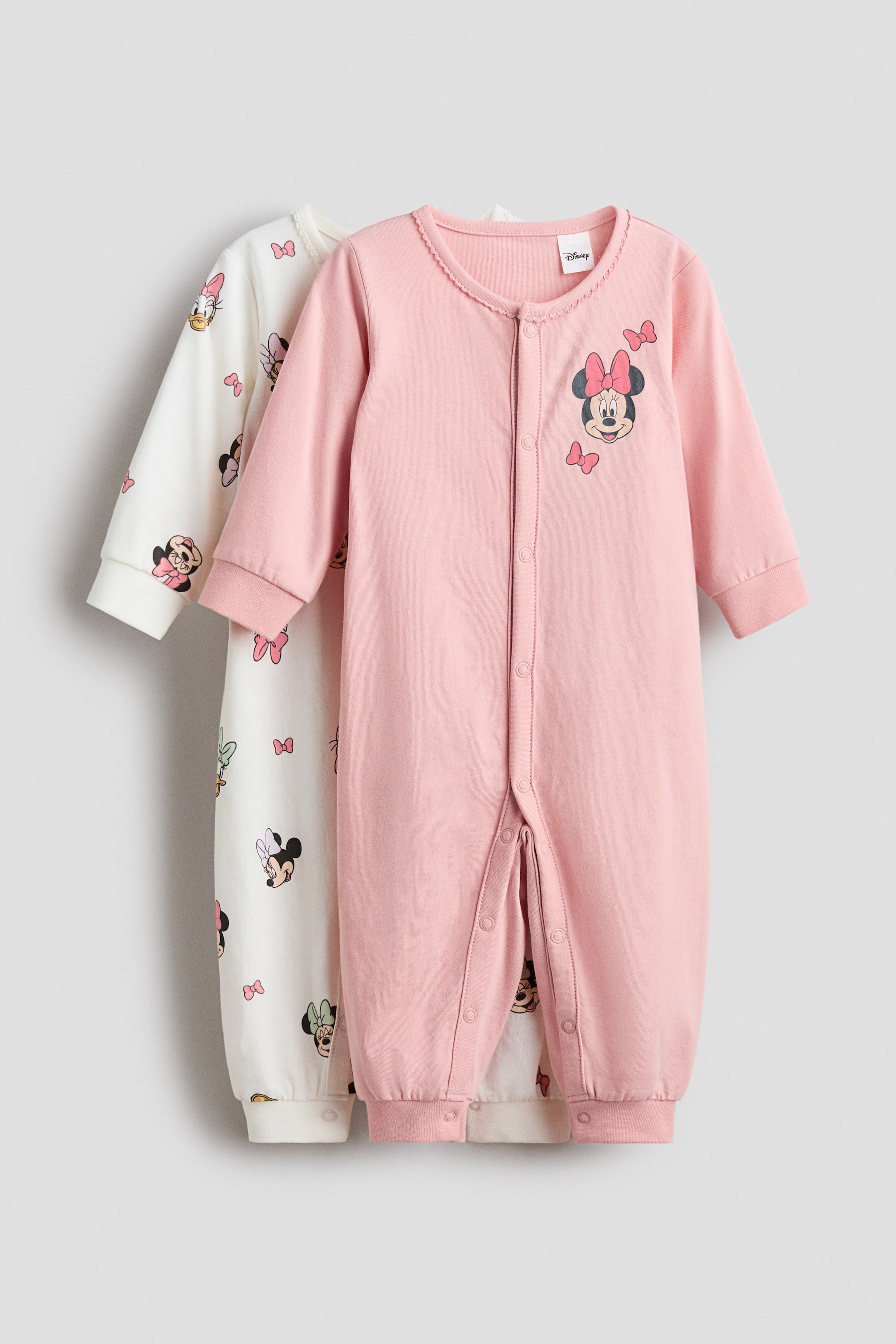 Xem ảnh lớn hơn: Hai bộ bodysuit bằng vải cotton in hình được hiển thị từ mặt trước, cả hai đều có tay dài, cổ tay và viền gân, và nút bấm kéo dài xuống phía trước và một chân. Bộ bodysuit màu hồng ở phía trước có hình ảnh Minnie Mouse trên ngực, trong khi bộ màu kem phía sau có họa tiết toàn thân của Minnie Mouse, Daisy Duck, và nơ.