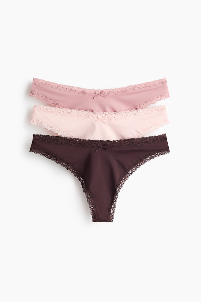 Drei Tanga-Slips liegen flach in einer gestapelten Anordnung und präsentieren ihre Vorderansicht. Der obere Slip ist in einem staubigen Rosa, der mittlere in einem hellen Rosa und der untere in einem dunklen Braun gehalten. Jeder Slip ist mit einem zarten Spitzenbesatz entlang der Taille und Beinöffnungen versehen und wird durch eine kleine Schleife am vorderen Bund verziert. - 1