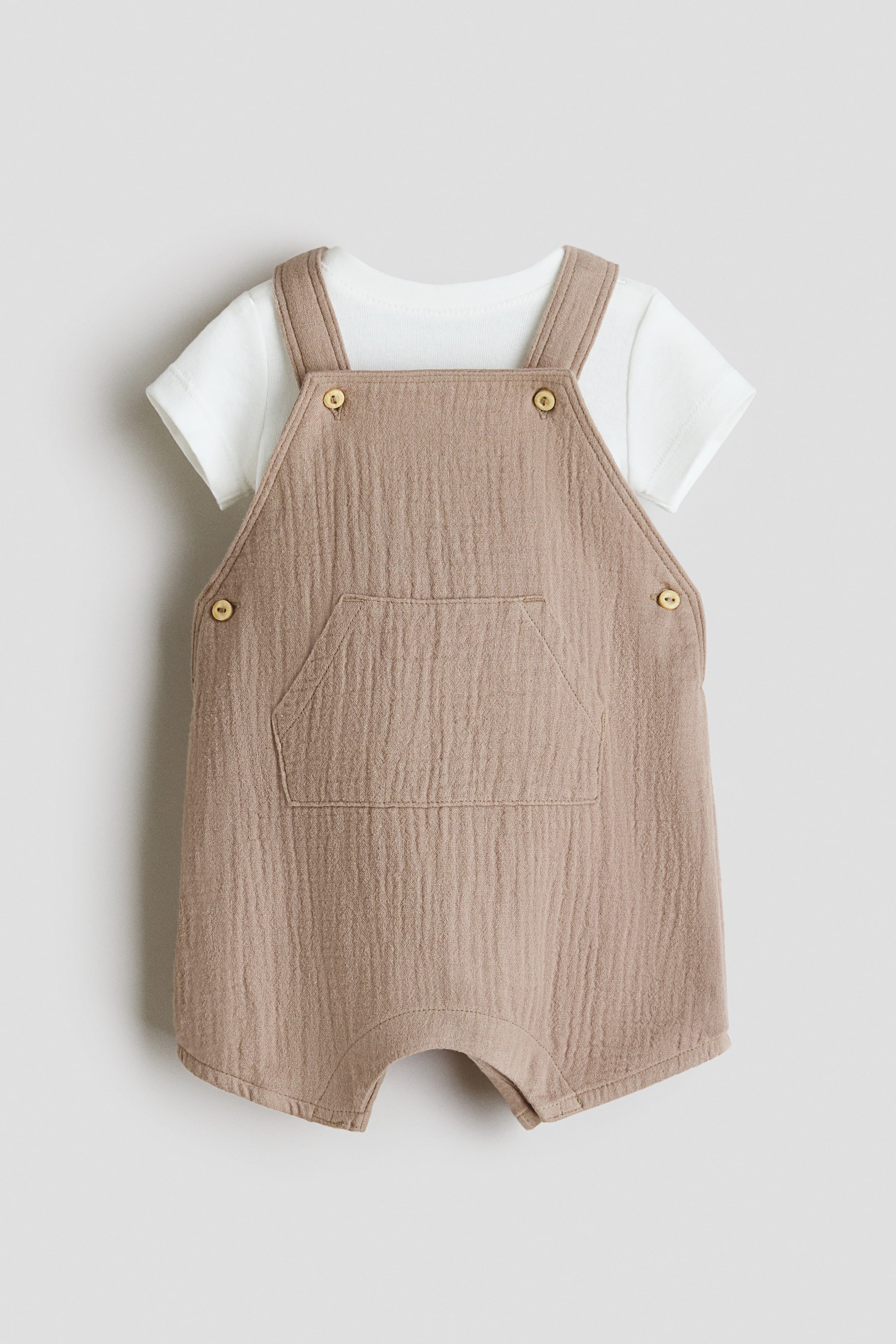 Grotere afbeelding bekijken: Een vooraanzicht toont een babyset, bestaande uit een wit ribgebreid katoenen jersey rompertje met korte mouwen, gedragen onder lichtbruine katoenen geweven tuinbroek. De tuinbroek heeft een kangoeroezak op het voorpand, verstelbare bandjes met knopen in houtlook en een knoopsluiting aan de linkerzijde.