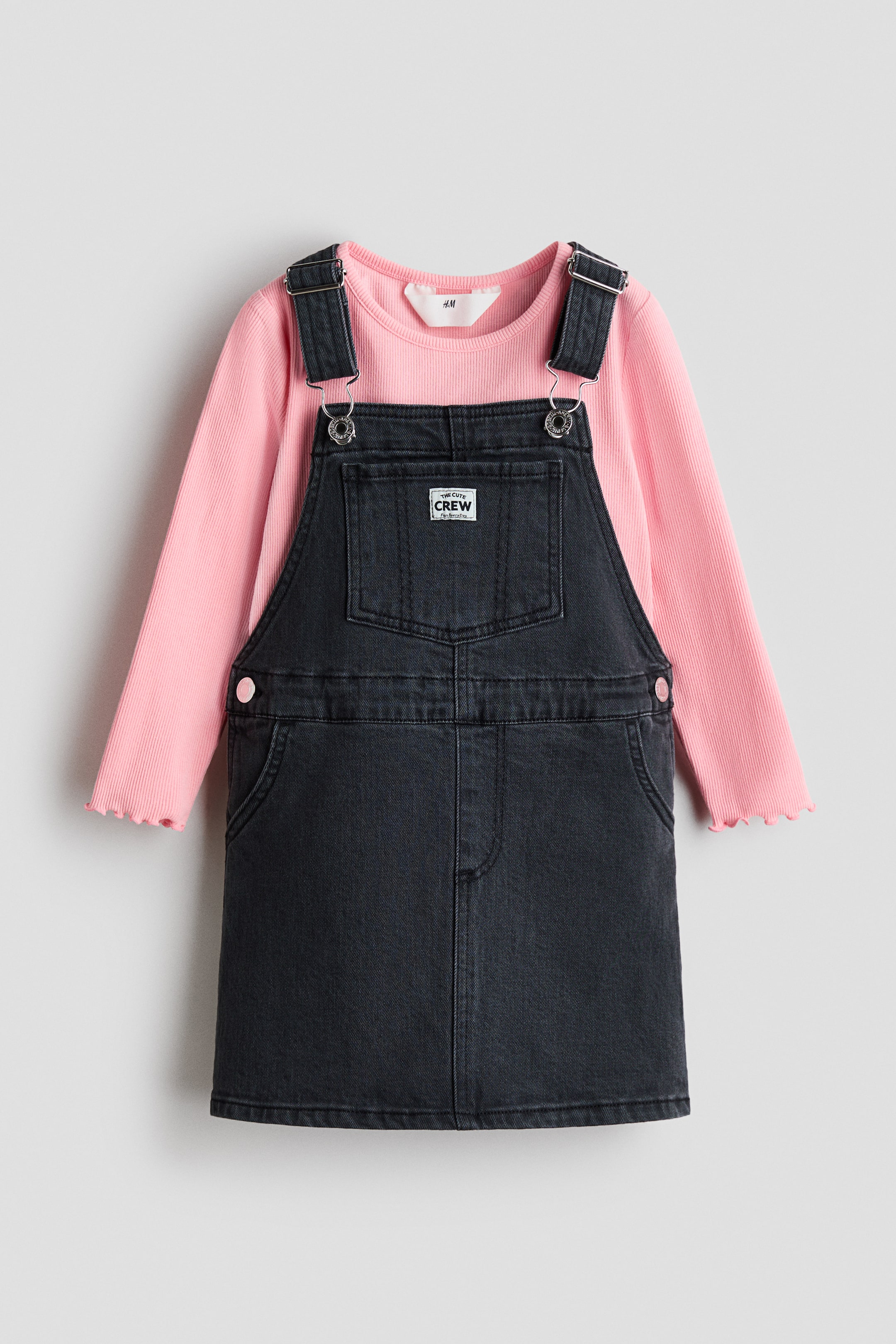 View larger image: A black denim overall dress is layered over a pink ribbed long-sleeved top, showcased from the front. The overall dress features adjustable shoulder straps with silver-tone buckles and a front bib pocket displaying a white "THE CUTE CREW" label. The pink top exhibits a crew neck and lettuce-edge cuffs.
