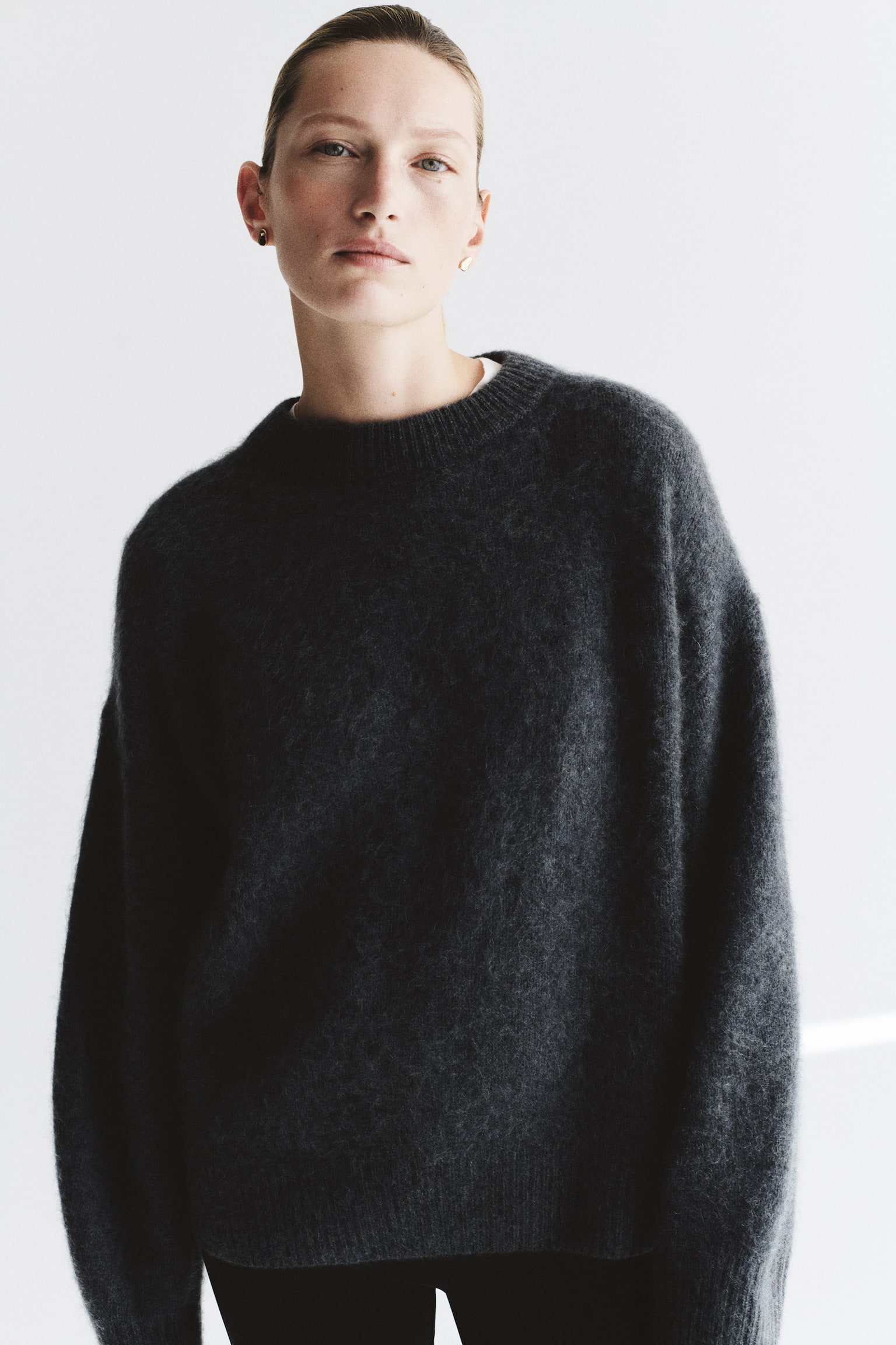 An oversized knitted jumper in dark grey is shown from the front, featuring a soft, textured wool finish. It is designed with a round, rib-trimmed neckline, dropped shoulders, and long sleeves with wide ribbed cuffs and a matching ribbed hem. - 1