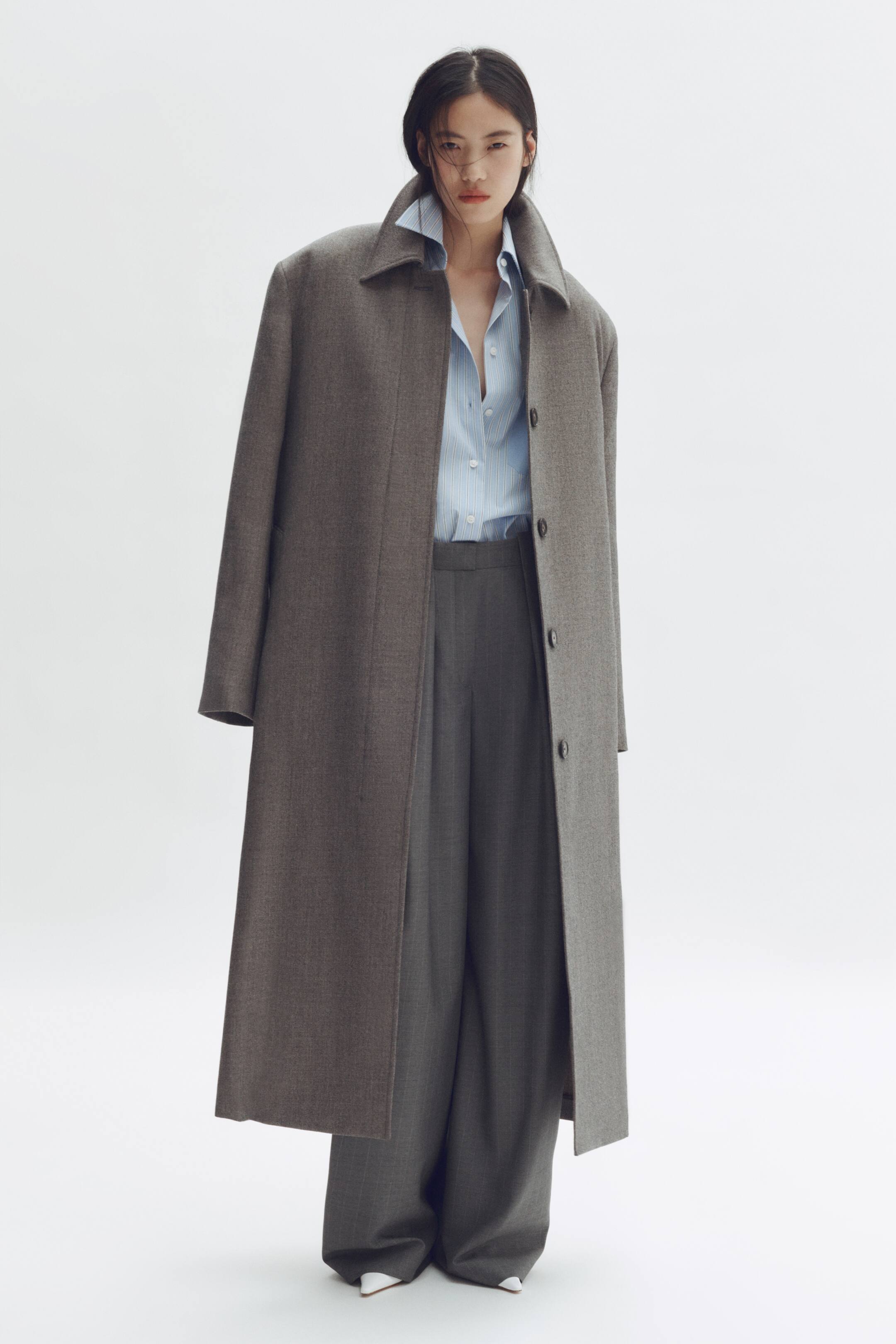 View larger image: A dark grey long coat is facing forward, featuring a pointed collar, notched lapels, and a single-breasted front with three visible buttons. It is being worn open over a light blue pinstriped shirt and dark grey wide-leg trousers, showcasing its oversized fit and full length.
